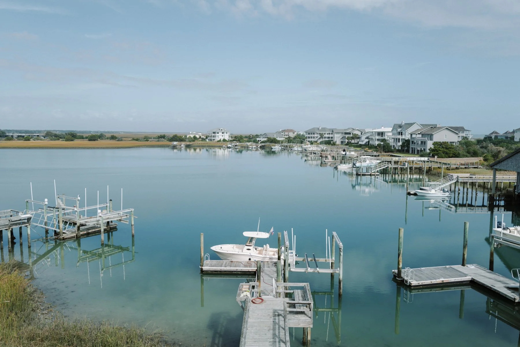 View of a calm marina with boats docked at wooden piers, surrounded by residential houses with balconies, under a partly cloudy sky.