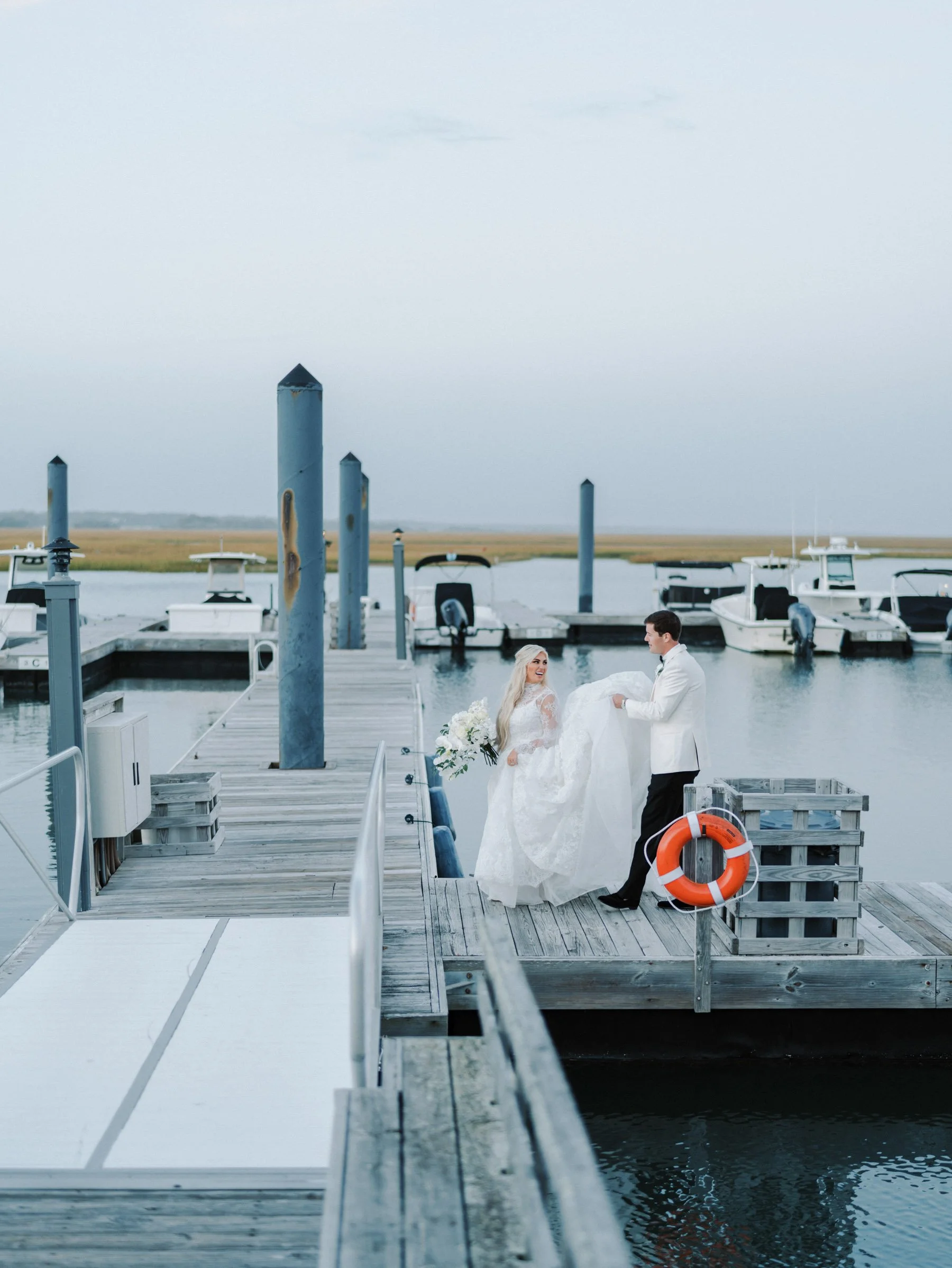A bride and groom at a dock by the water, with boats in the background. The groom is lifting the bride in her wedding dress, smiling at her.