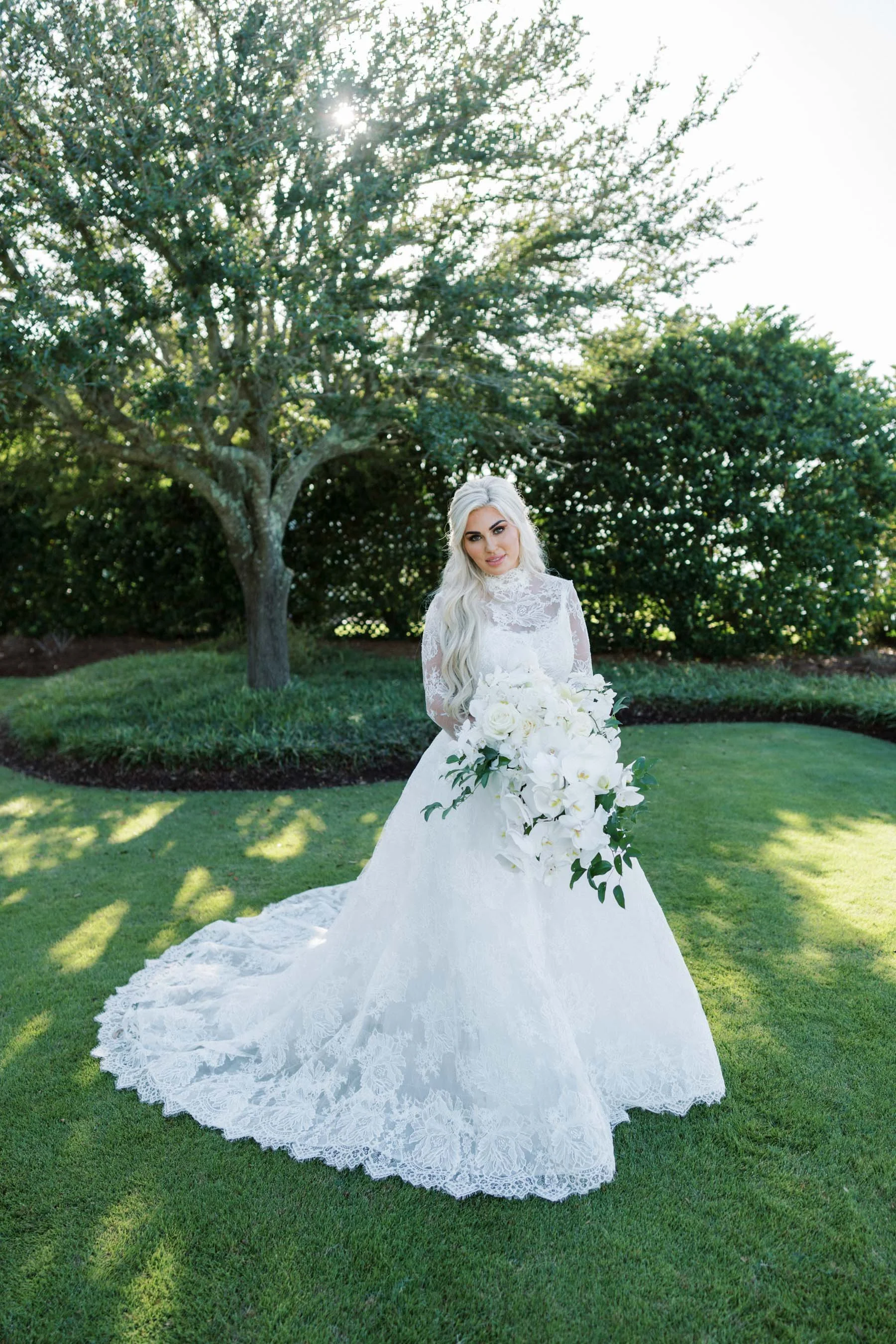A bride in a white lace wedding dress holding a large bouquet of white flowers, standing on a well-manicured green lawn with trees and bushes in the background, under a sunny sky.