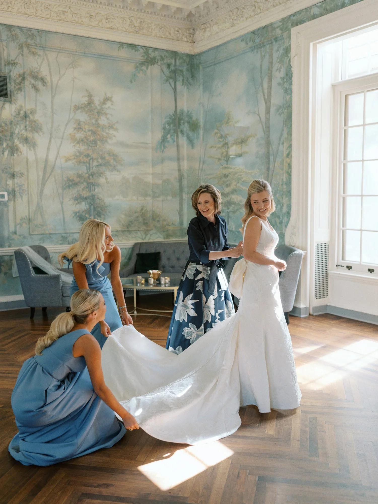 A bride getting ready for her wedding with her bridesmaids and possibly her mother helping her with her wedding dress in an elegant room with large windows and a detailed mural on the wall at the Curtis Arboretum.
