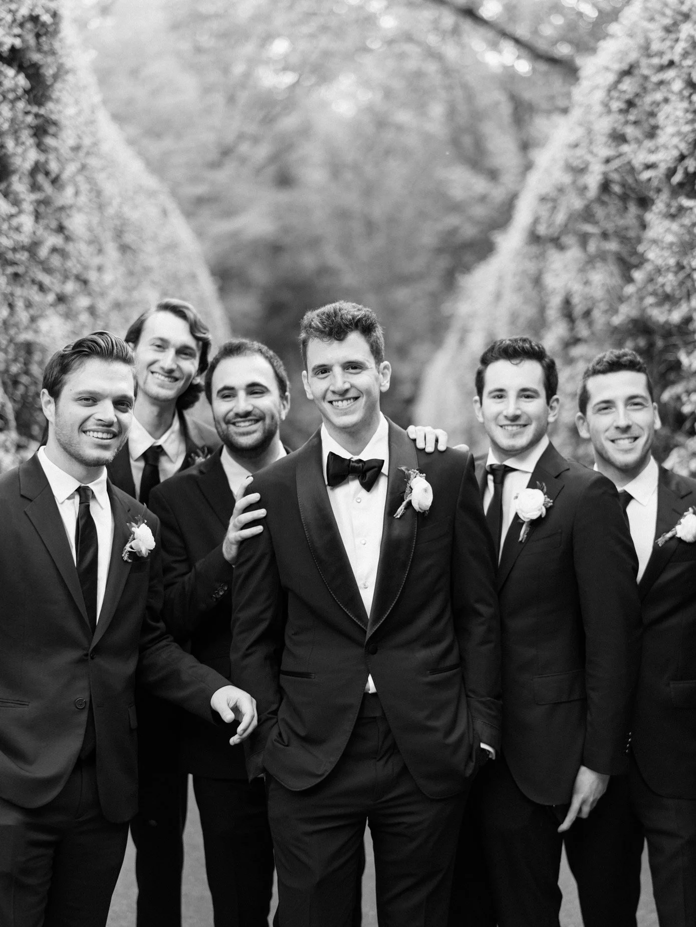 Group of six men dressed in tuxedos, smiling for a photo outdoors, possibly at a wedding or formal event.