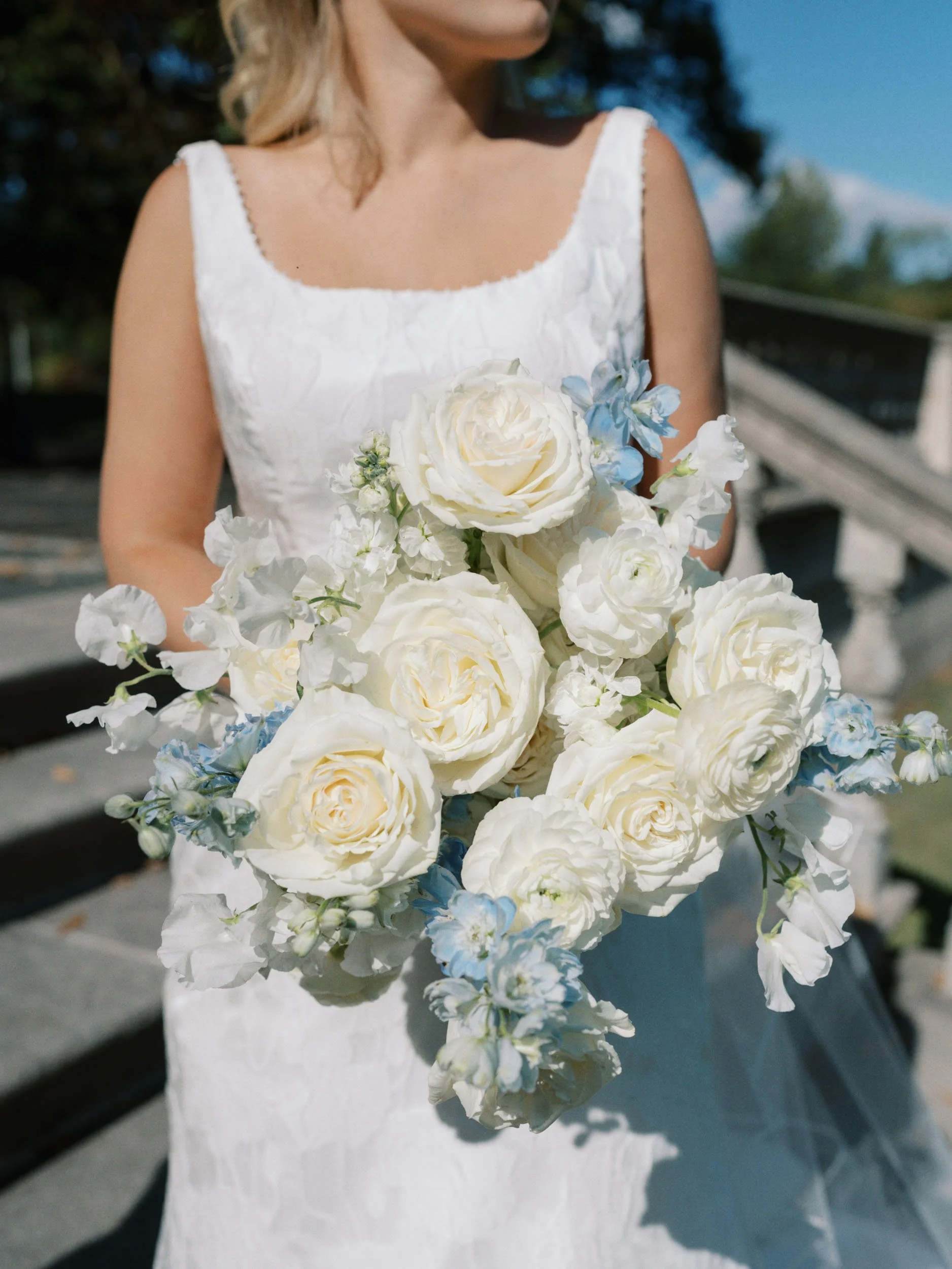 A woman in a white dress holding a bouquet of white roses with some light blue flowers outdoors on a sunny day.