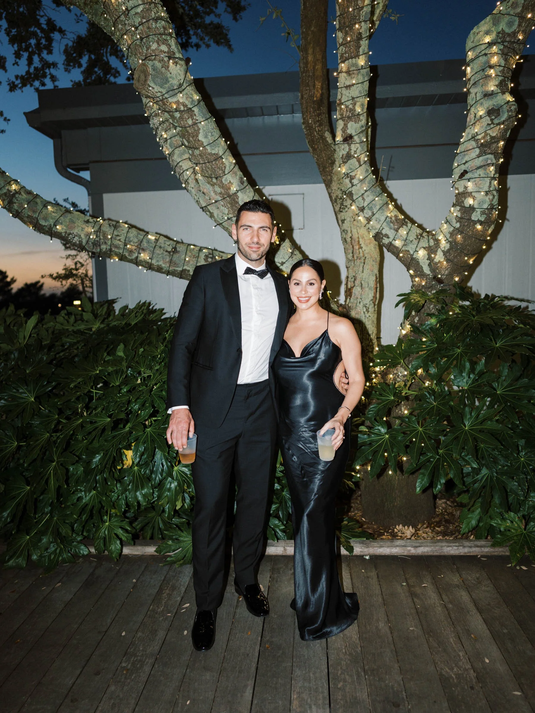 A man and woman dressed in formal evening wear standing outdoors during the evening, holding drinks, with a tree wrapped in string lights behind them.