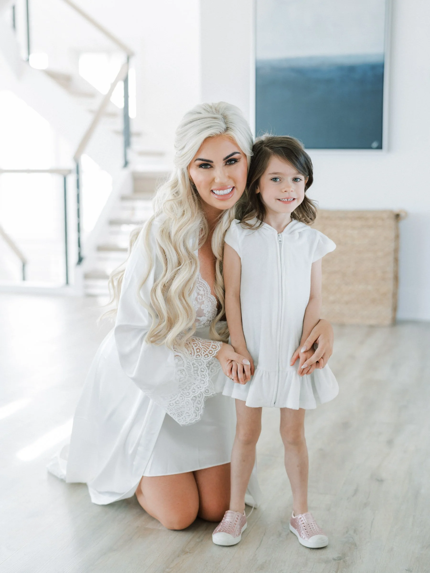 A woman with long blonde hair kneeling and smiling, holding hands with a young girl with brown hair, inside a modern, well-lit room.