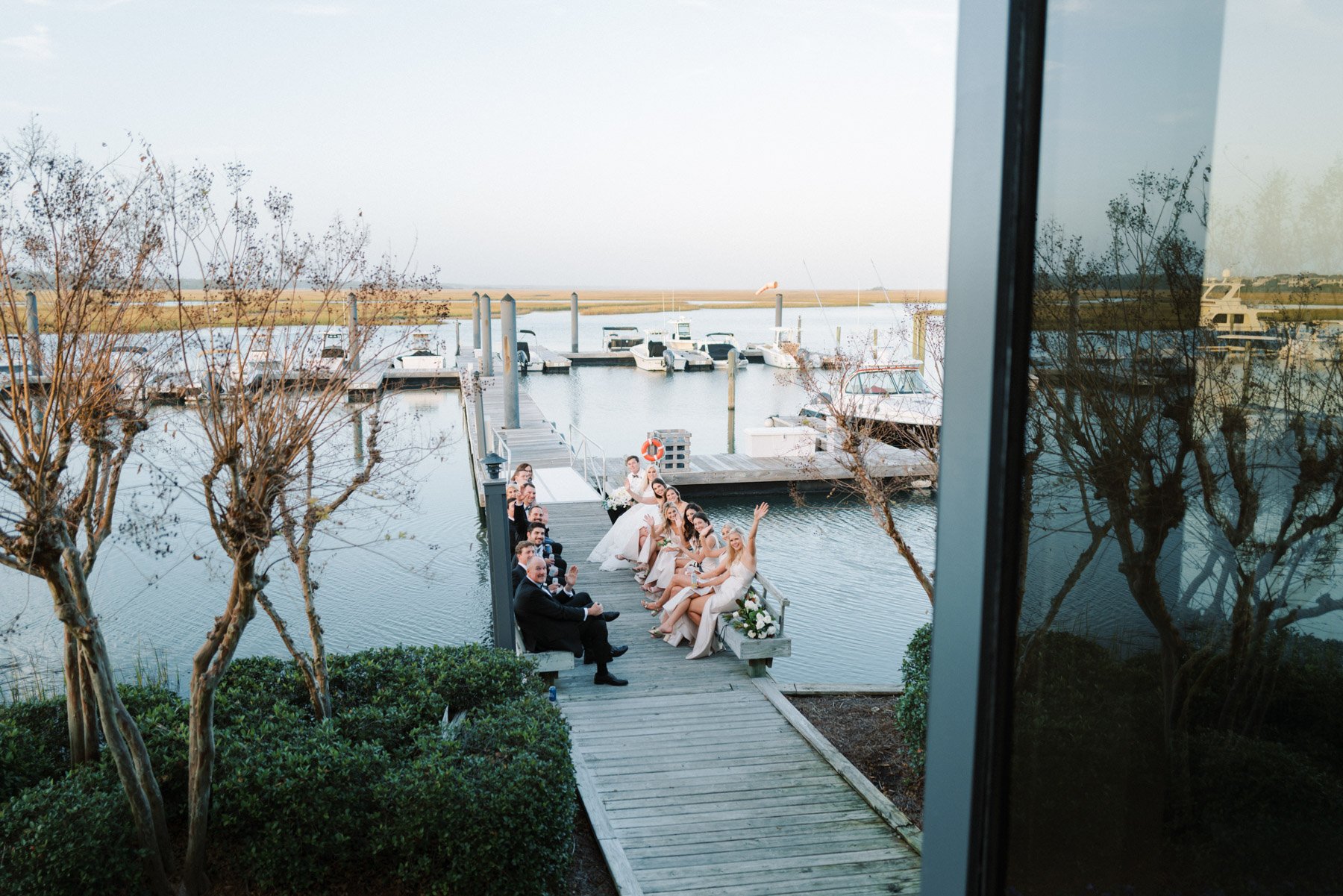 A group of people, including a bride in a wedding dress, are sitting on a dock beside a marina with boats, seen through a window, celebrating outdoors during daylight.