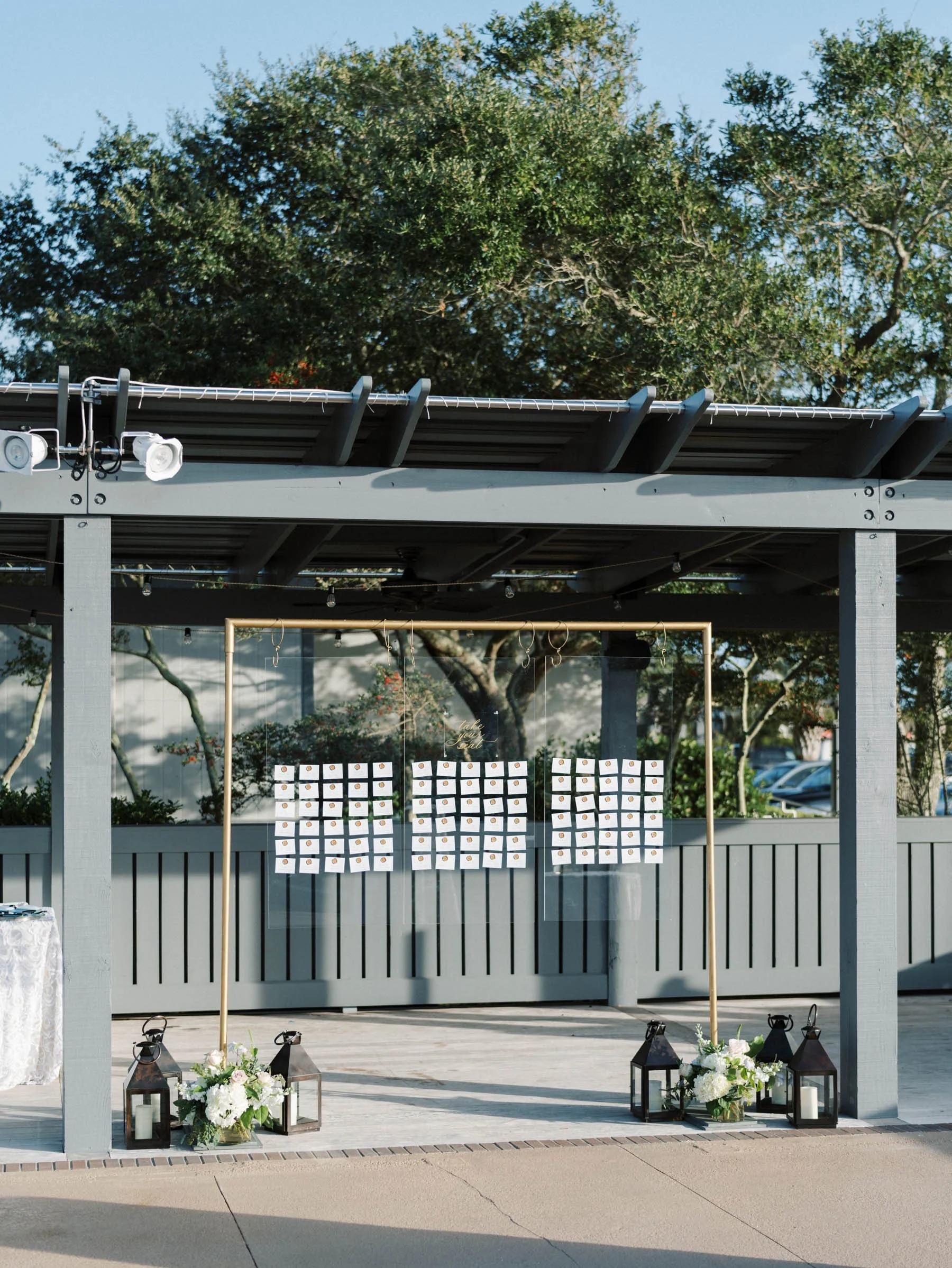 Outdoor event setup with a decorative gold frame holding rows of small cards, surrounded by lanterns and floral arrangements on the ground, under a gray wooden structure with string lights and surveillance cameras, with trees and parking lot in backg