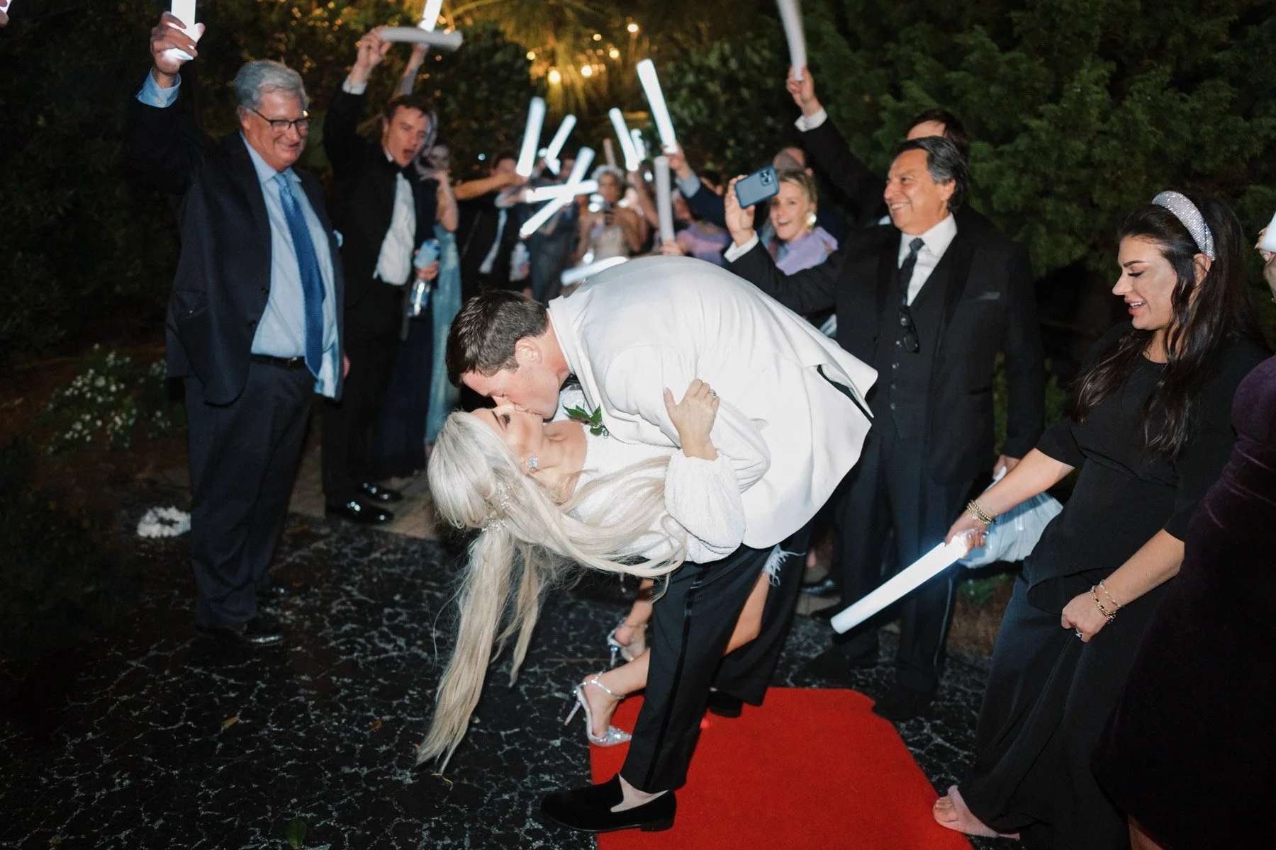 A wedding celebration outdoors at night with a bride and groom kissing, surrounded by guests cheering and taking photos, with some holding light-up items.
