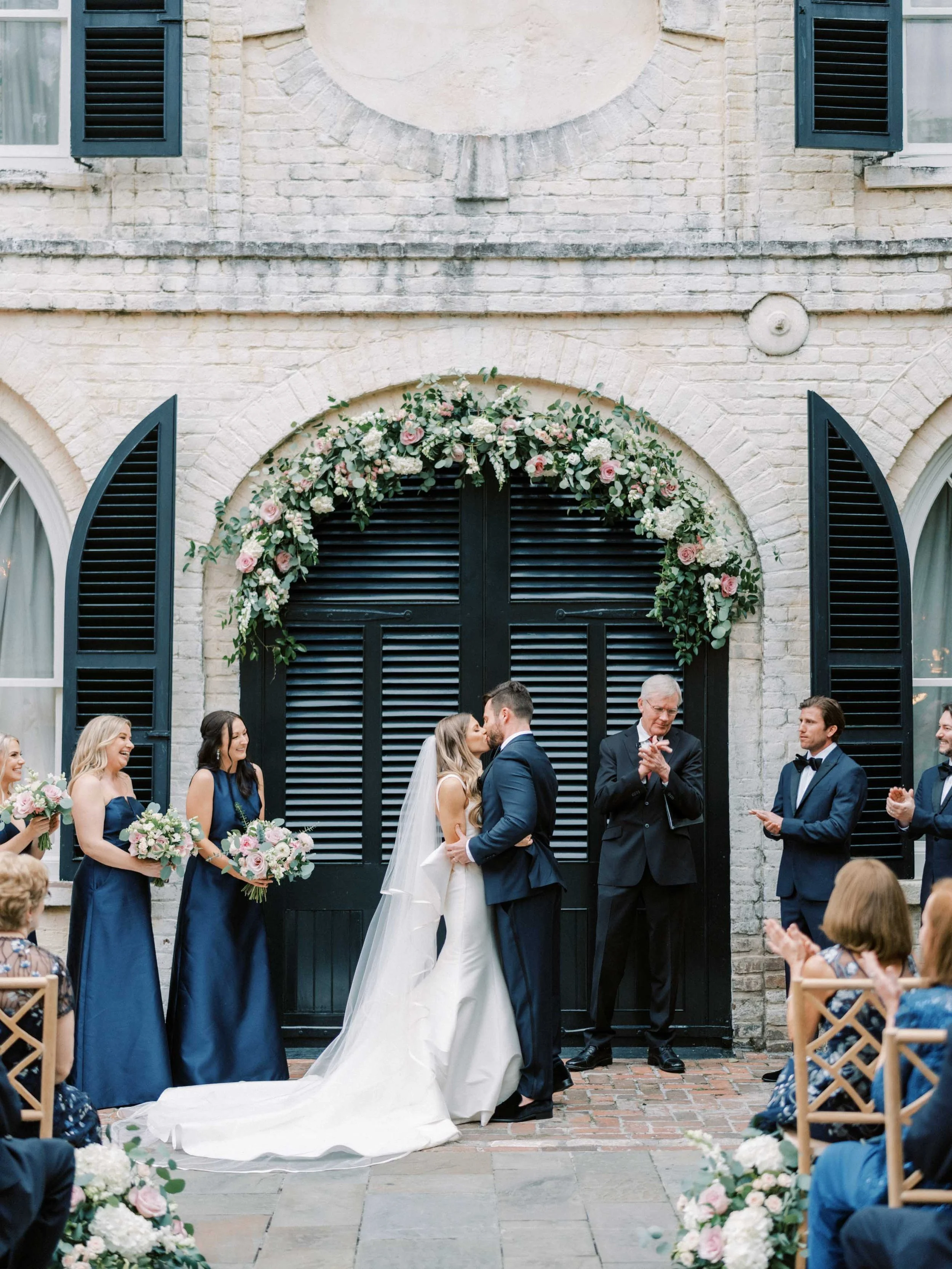 Bride and groom kissing during their wedding ceremony outdoors, with bridesmaids and groomsmen on either side, and guests seated around them.