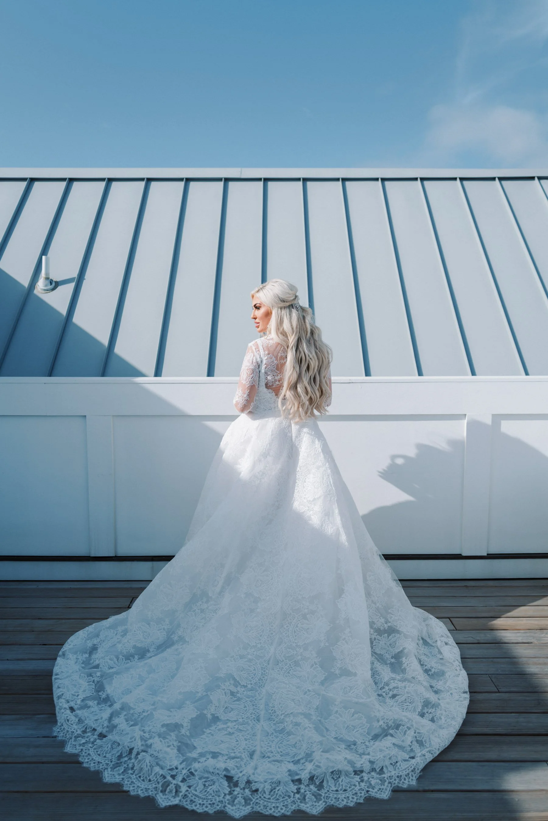 Bride in a white wedding gown with lace details, standing on a wooden deck against a white wall and a clear blue sky.