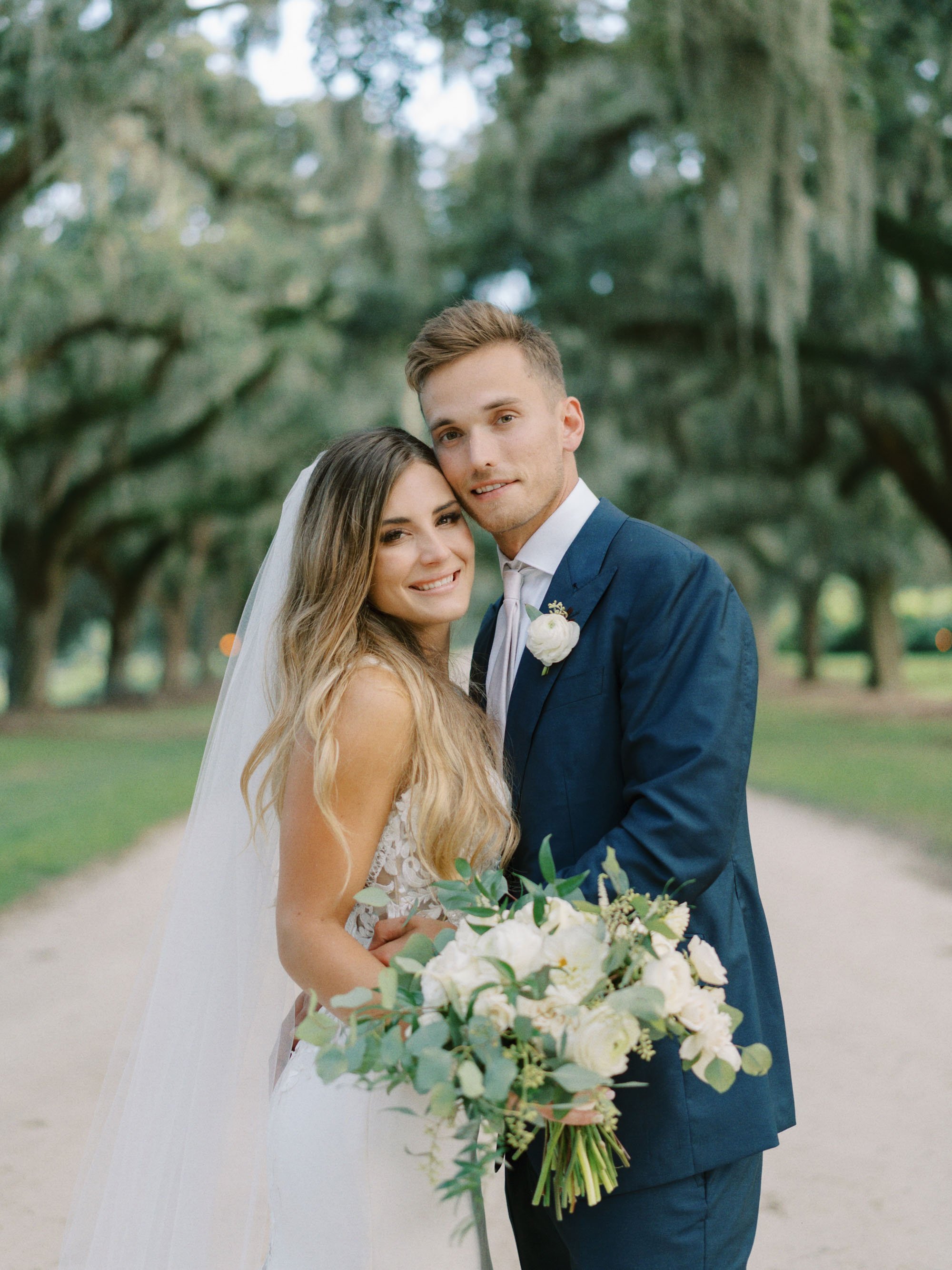 A newlywed couple standing outdoors on The Avenue of Oaks, with the bride holding a large bouquet of white flowers and the groom wearing a blue suit with a white boutonniere, surrounded by large trees and greenery in the background.