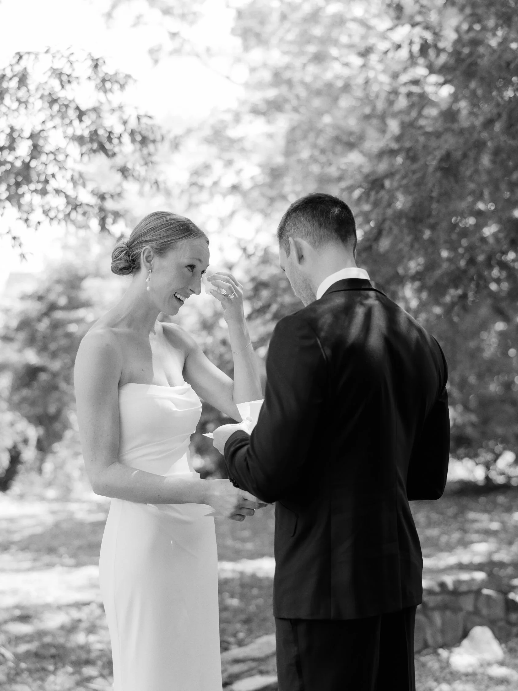A bride and groom exchanging vows outdoors, with the bride smiling and wiping a tear, surrounded by trees.