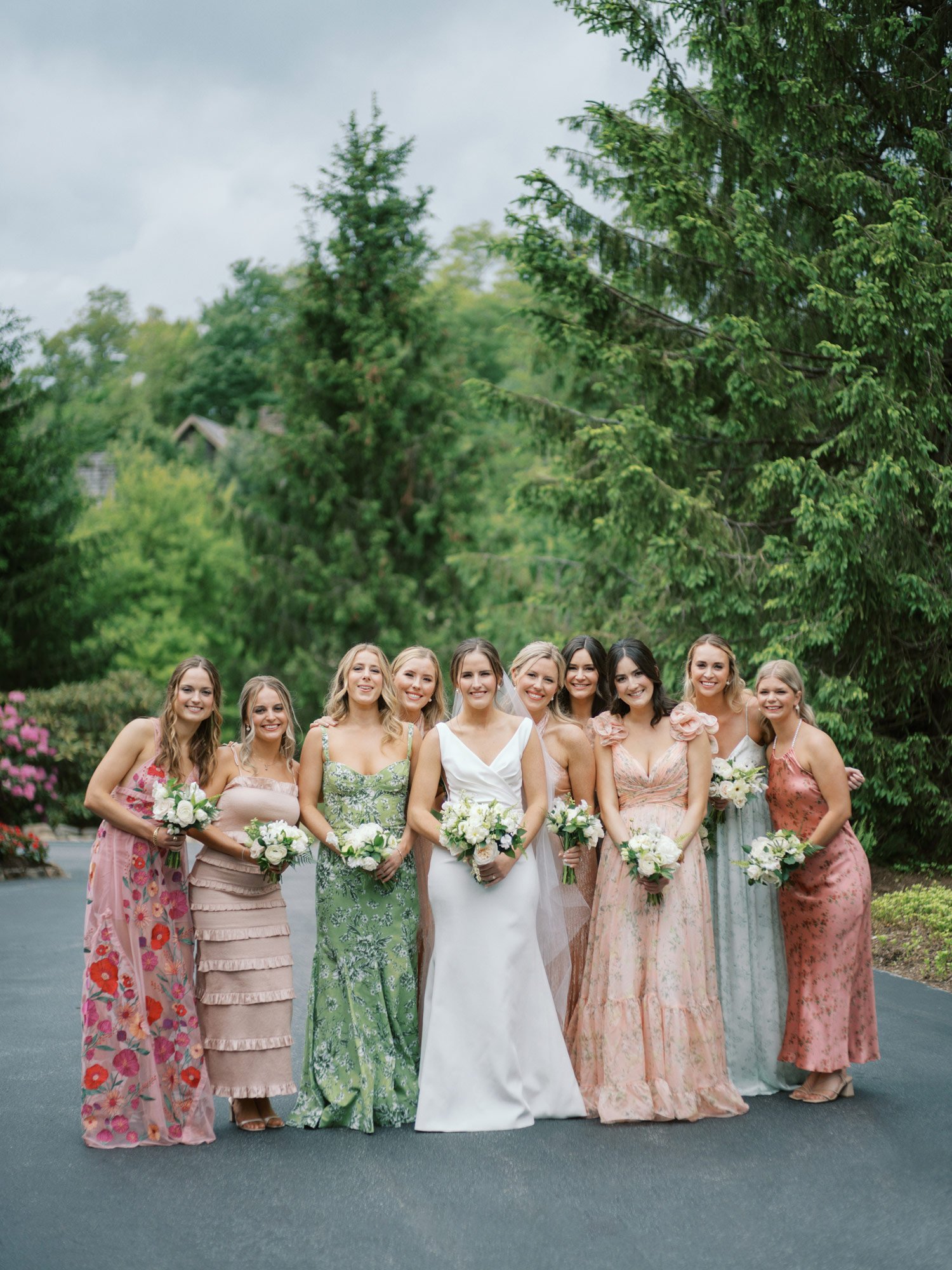 A group of nine women, including a bride in a white gown, stand together outdoors on a paved area with green trees behind them. They are holding white flower bouquets and wearing colorful dresses.