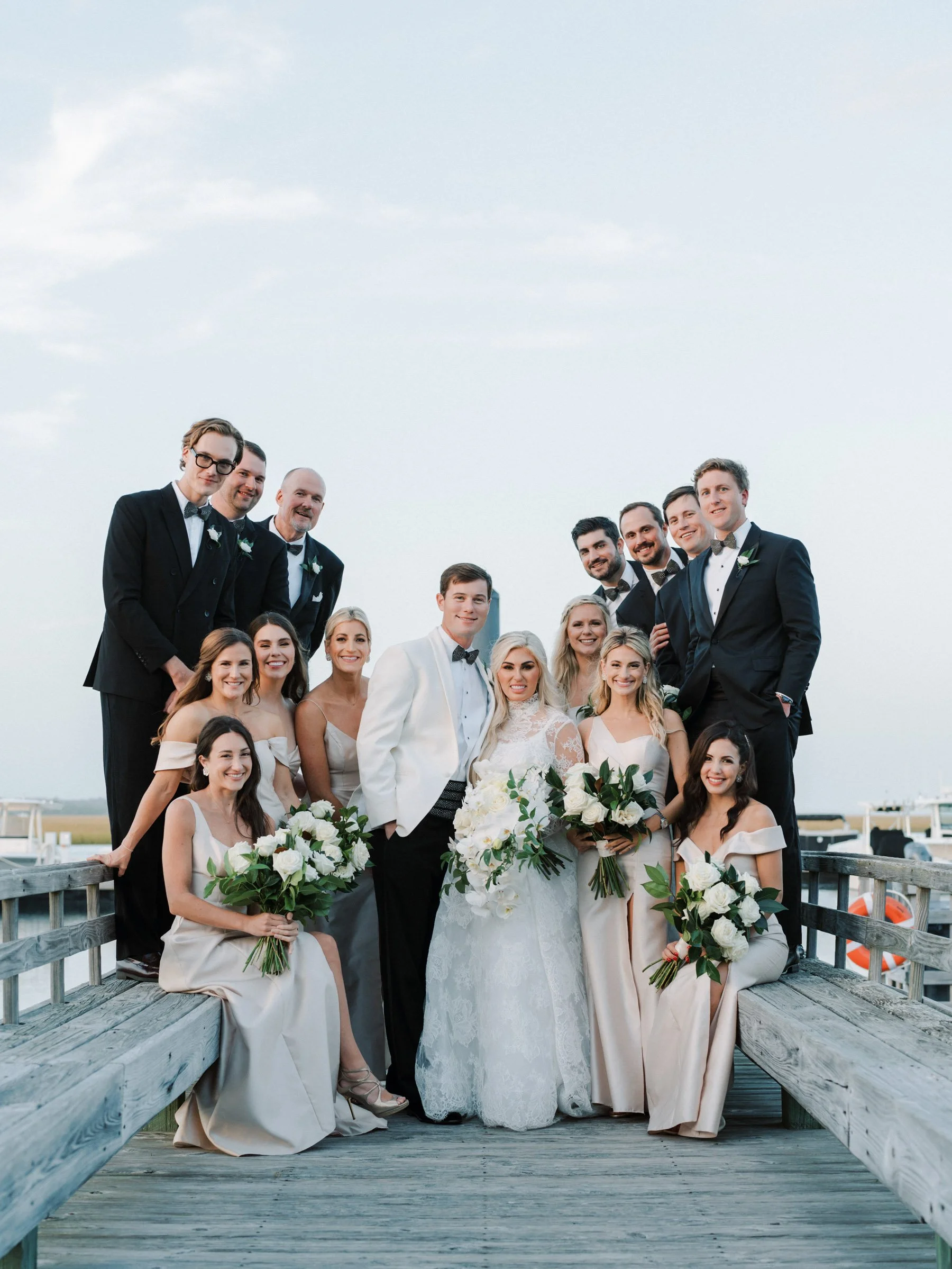 A wedding group photo of the bride, groom, bridesmaids, and groomsmen on a wooden pier outdoors with a cloudy sky in the background.