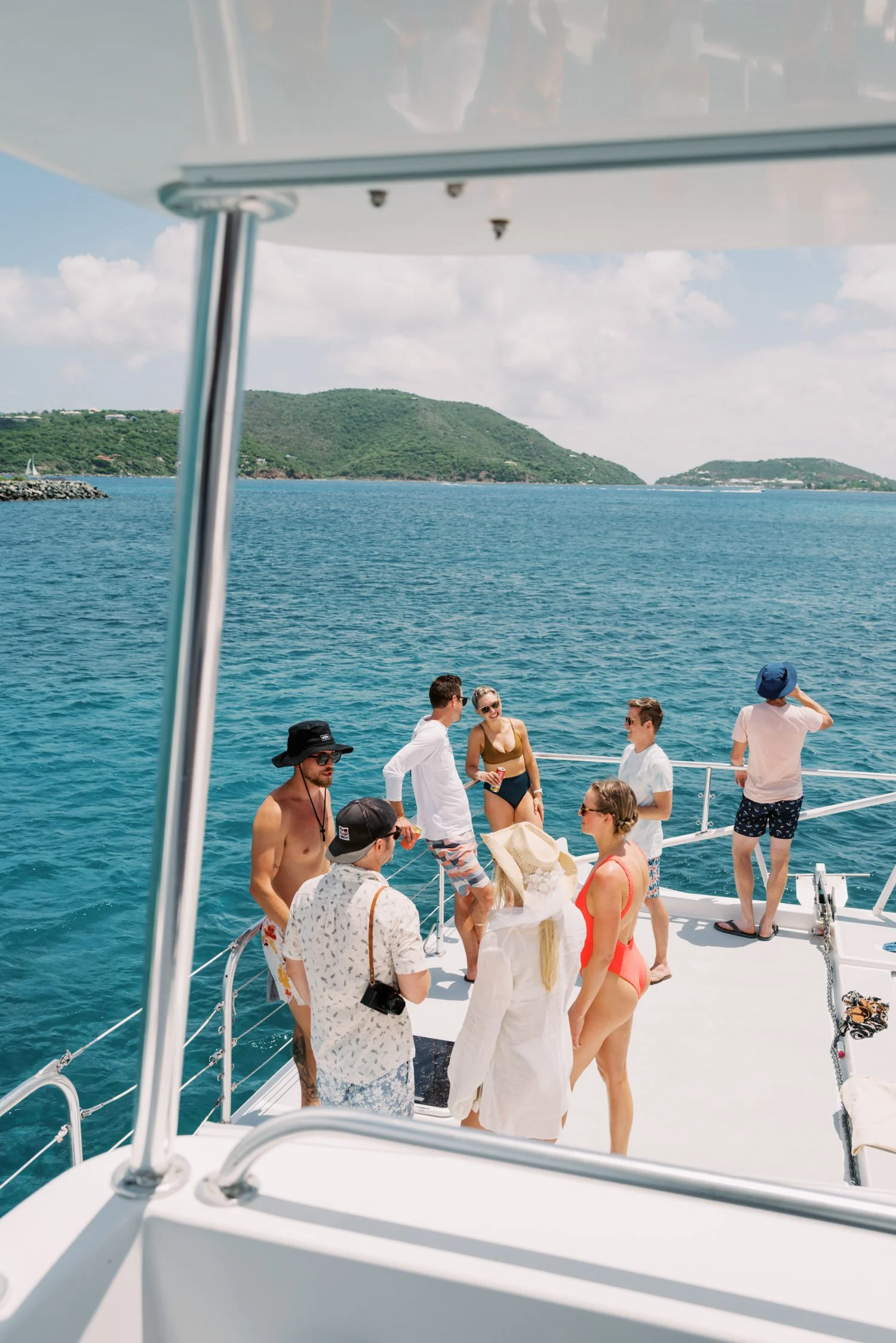 Group of friends on a boat enjoying a day in the water, with some wearing swimsuits and others in casual summer clothing, and a scenic view of islands and open sea in the background.