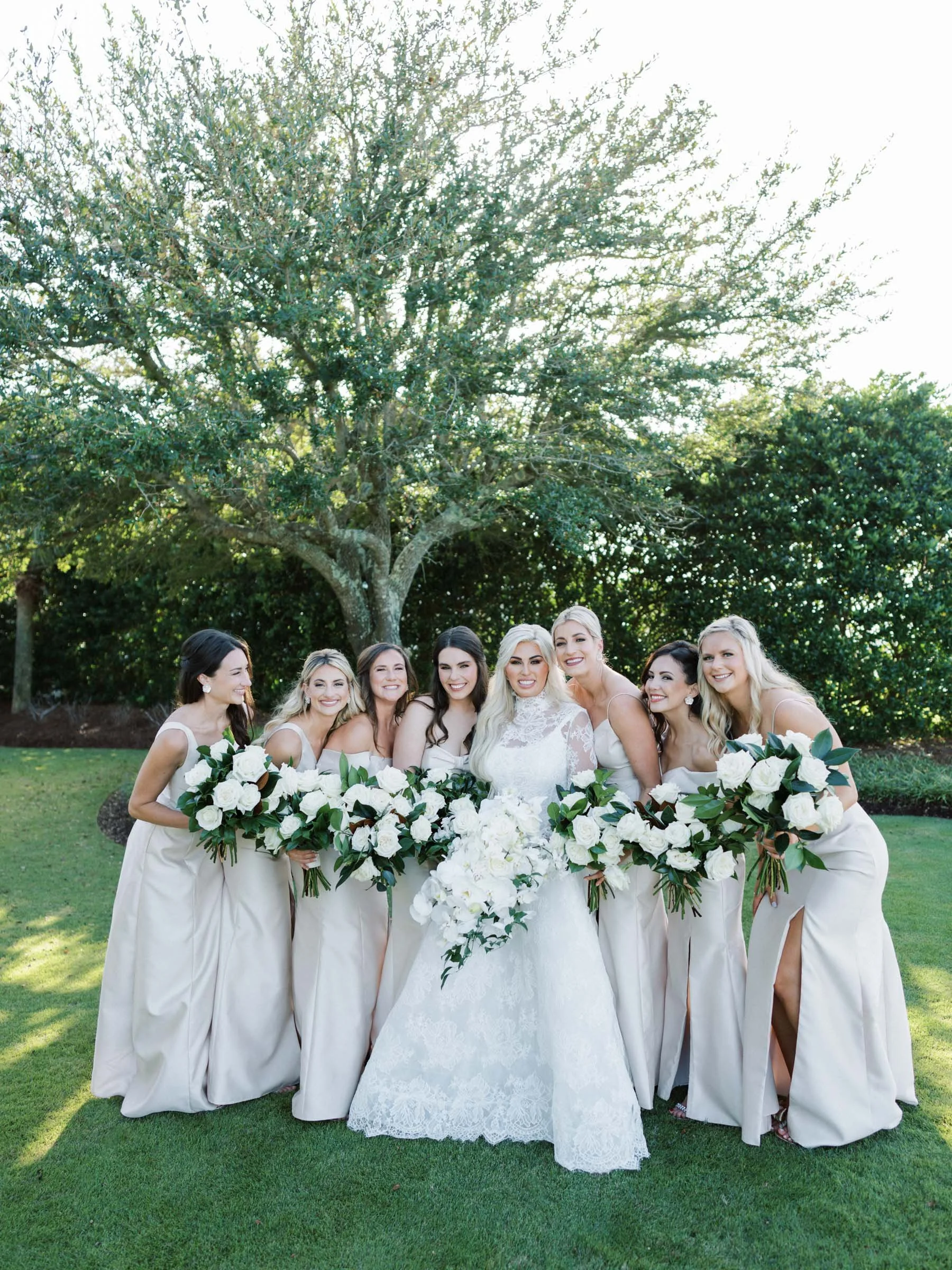 A bride in a white wedding dress with lace detail and six bridesmaids in matching long, off-white dresses, all holding large floral bouquets of white flowers and greenery, outdoors on a grassy area with trees in the background.