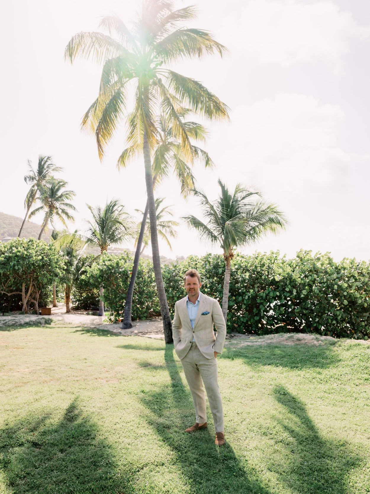 A man in a beige suit stands on a grassy area with palm trees and green bushes in the background, under a bright sunny sky.