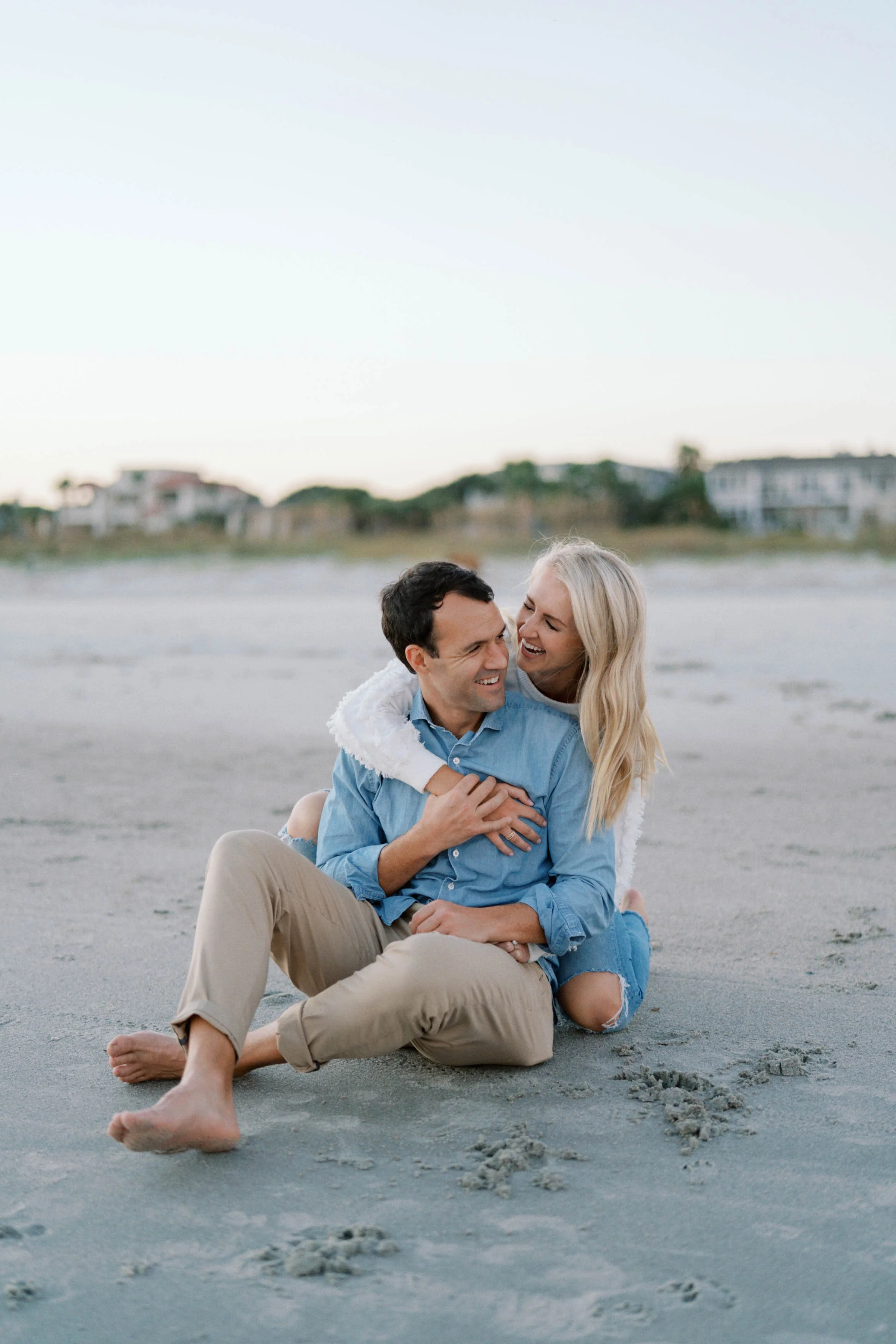 A happy couple sitting on the beach, embracing and smiling at each other, with houses in the background during sunset or late afternoon.