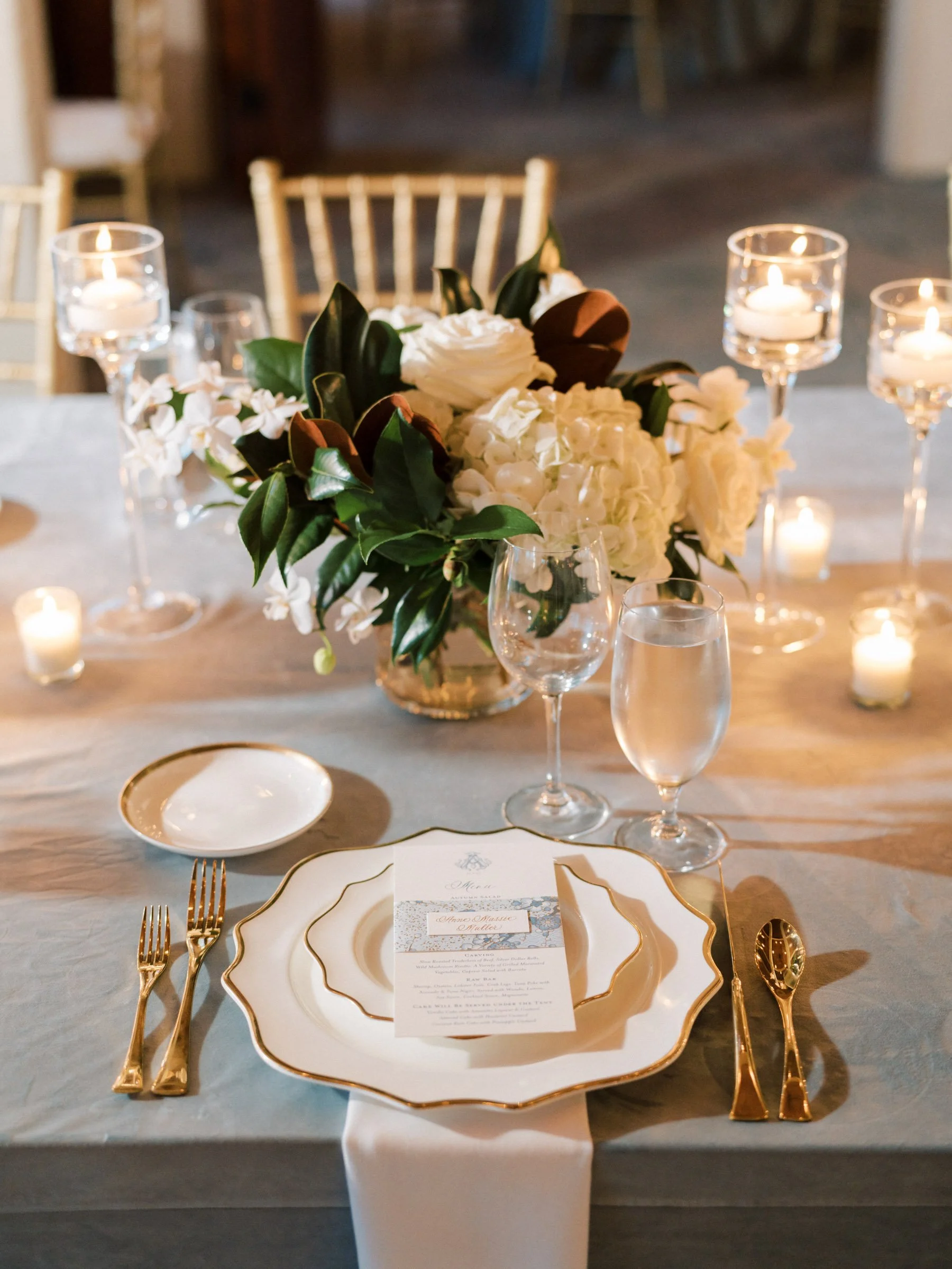 Elegant table setting with gold-edged plates, gold flatware, and a floral centerpiece of white flowers and green leaves, surrounded by lit candles.