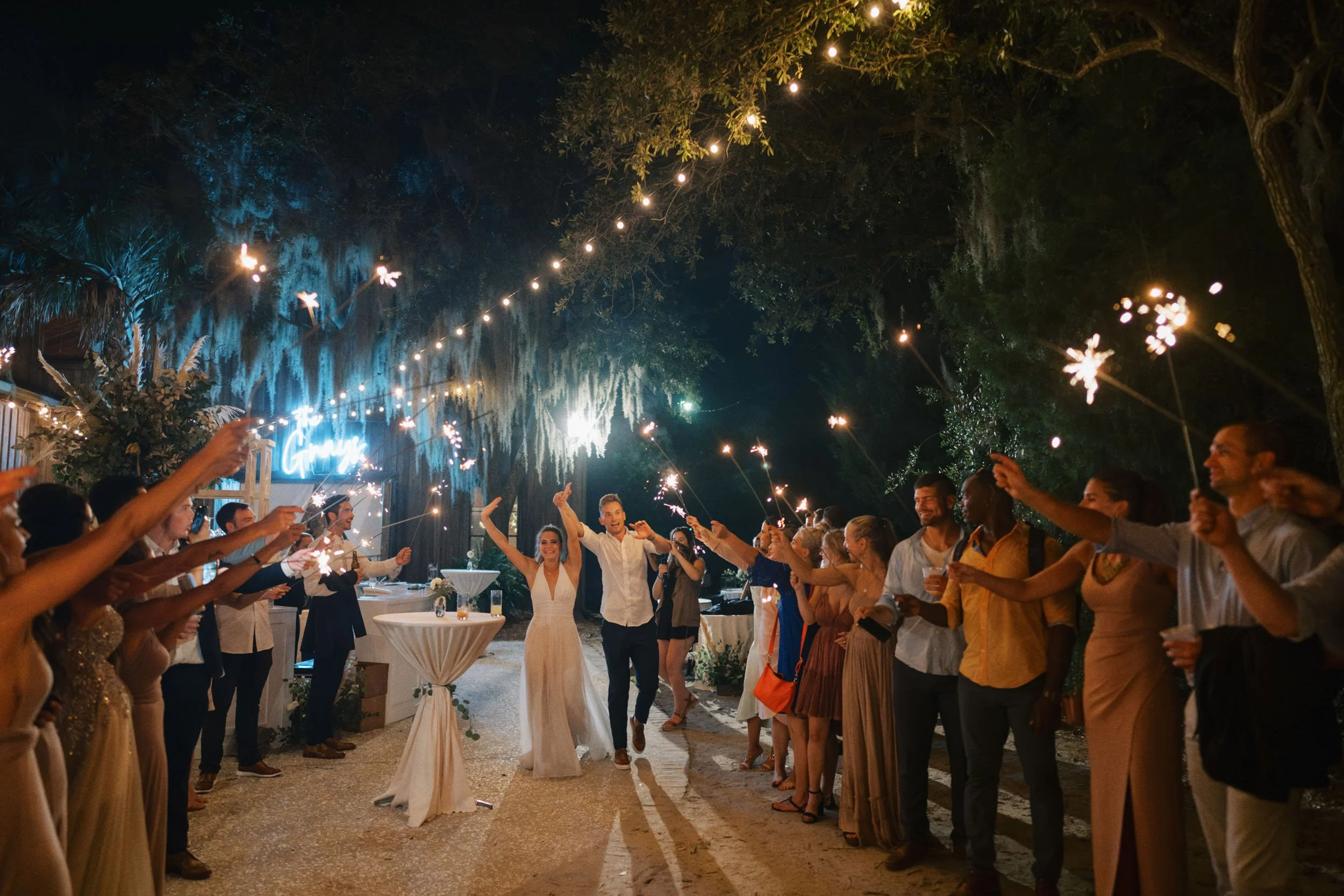 A couple in wedding attire walking through a celebration with friends holding sparklers at night, outdoors with trees and string lights.