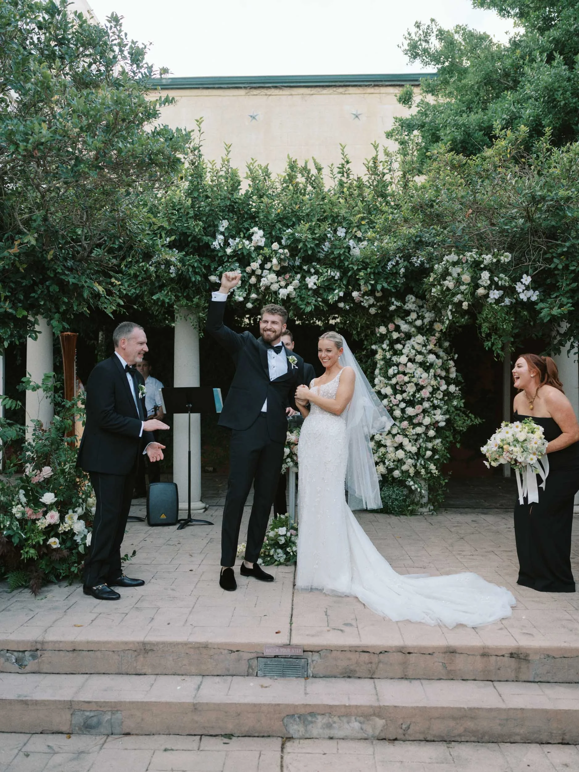 A wedding ceremony outdoors with the bride and groom celebrating with friends and family. The groom is raising his fist in joy, and the bride is smiling, holding his hand. A woman holding a bouquet stands to the right, smiling. The background features greenery and floral decorations.