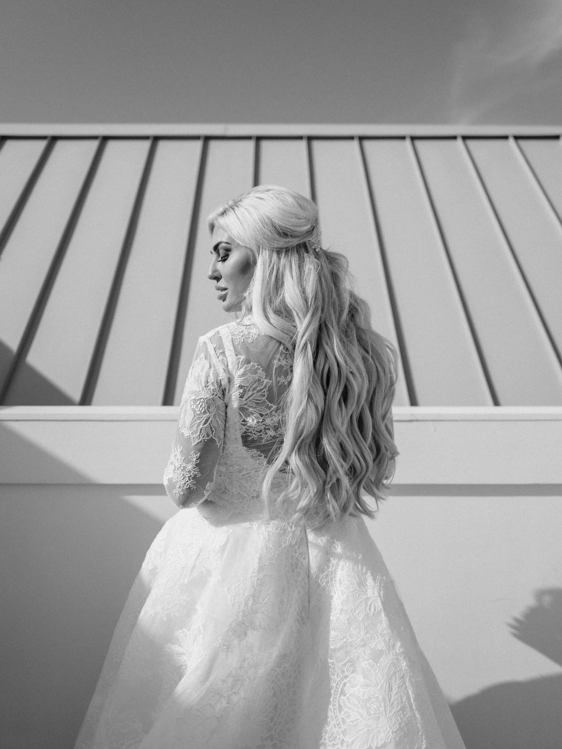 Black and white photo of a woman with long wavy hair in a lace dress, standing outdoors against a modern building with vertical bars.