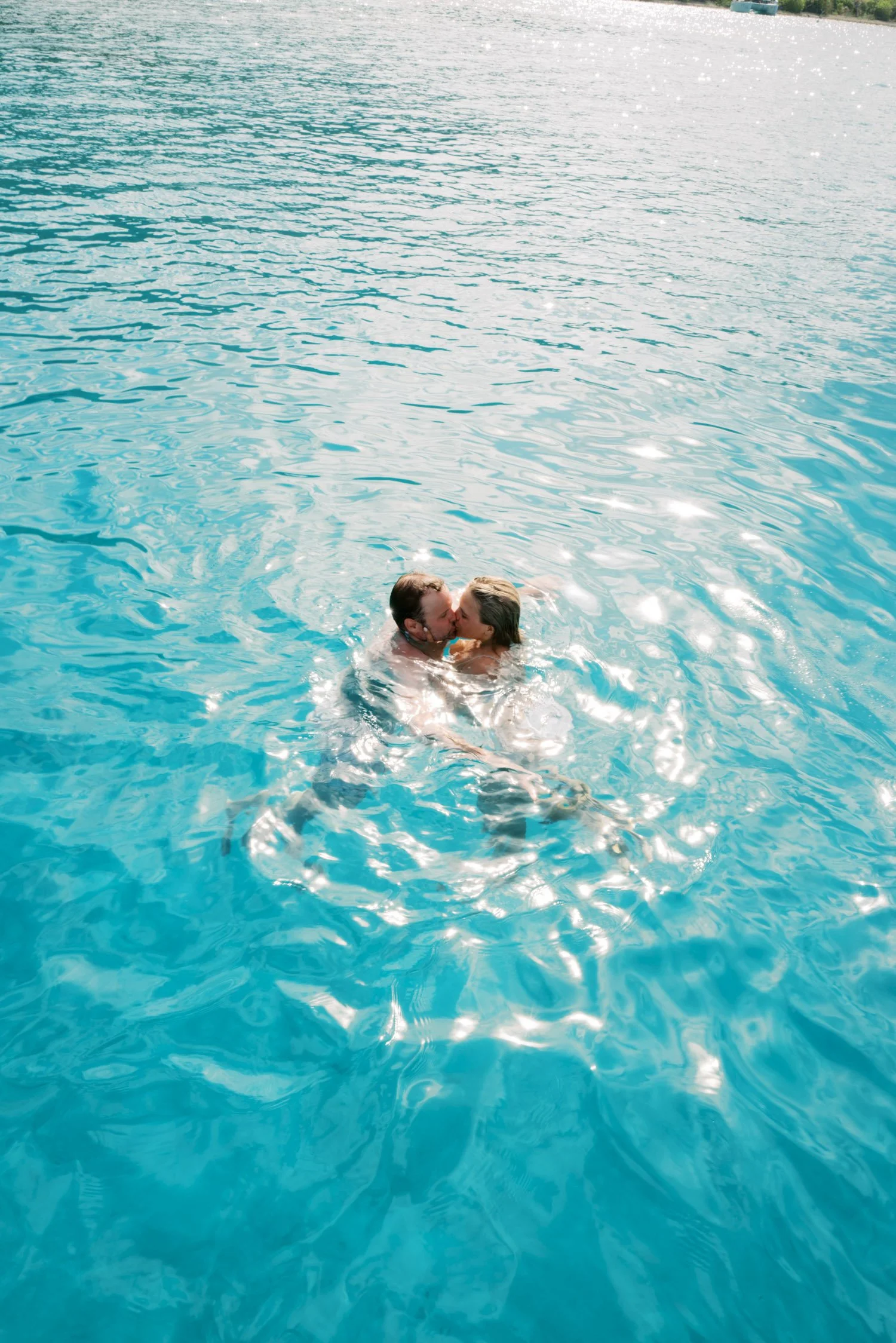 A man and woman sharing a kiss while swimming in a clear blue body of water, with sunlight reflecting on the surface.