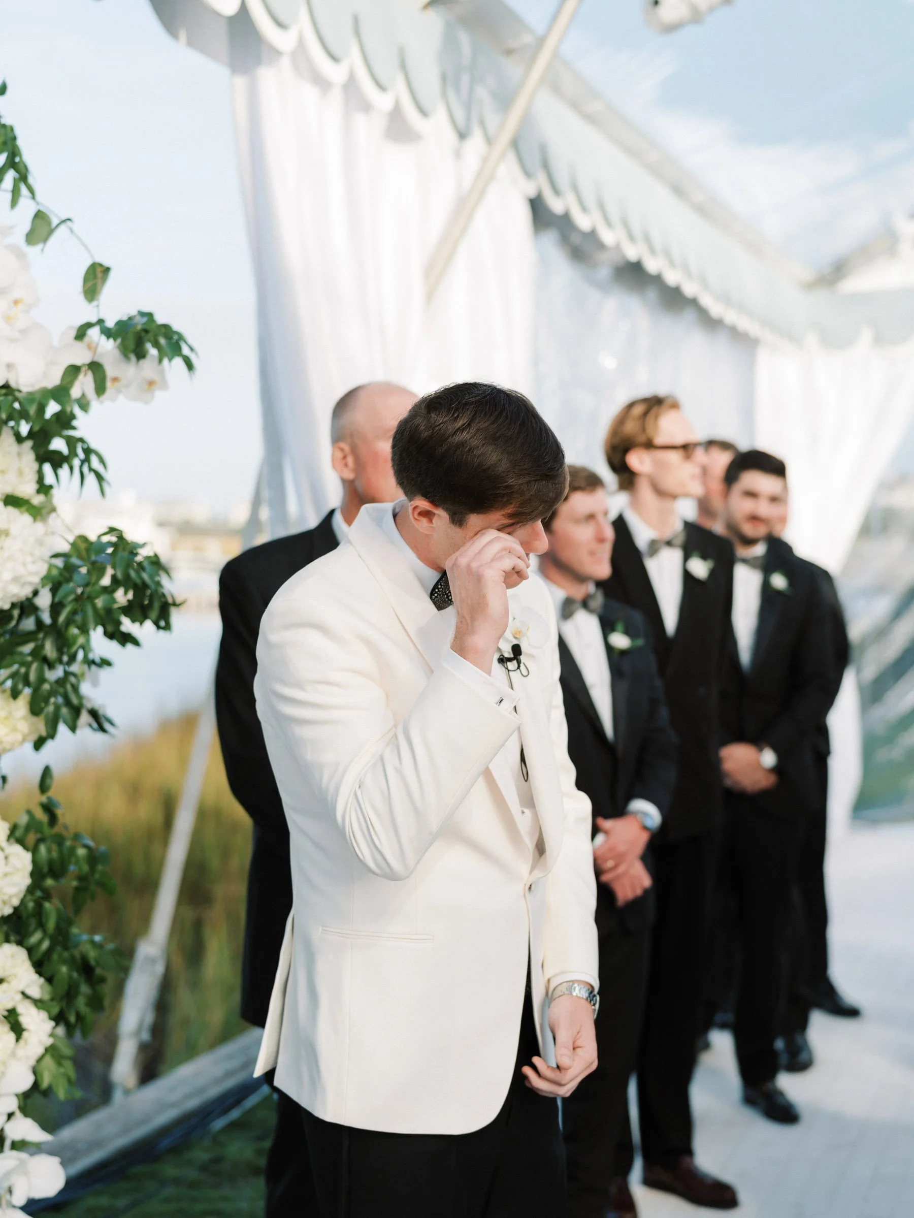A groom in a white tuxedo with a bow tie is emotional, wiping tears away, during a wedding ceremony. Behind him, five groomsmen in black tuxedos with bow ties stand under a draped, outdoor wedding arch decorated with white flowers.