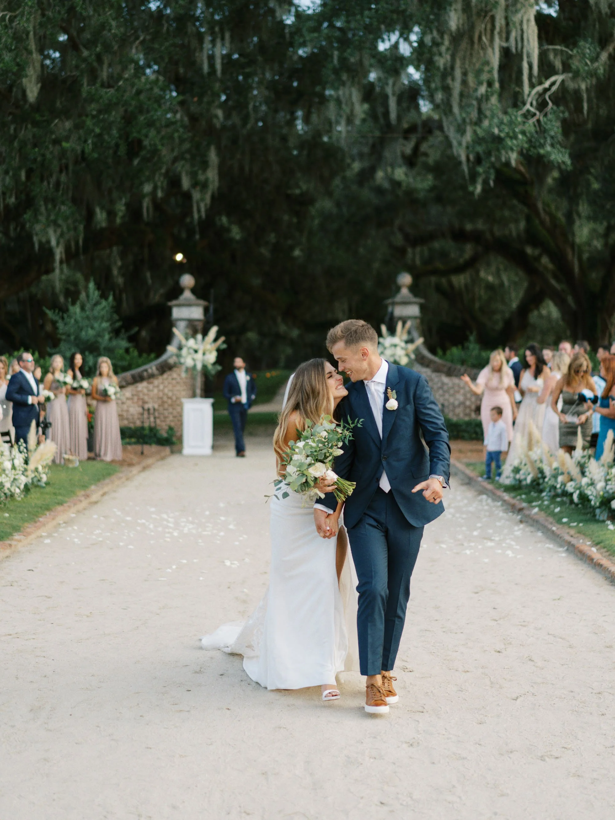 A bride and groom walk down the aisle at their outdoor wedding, surrounded by guests and floral decorations, under a large tree.