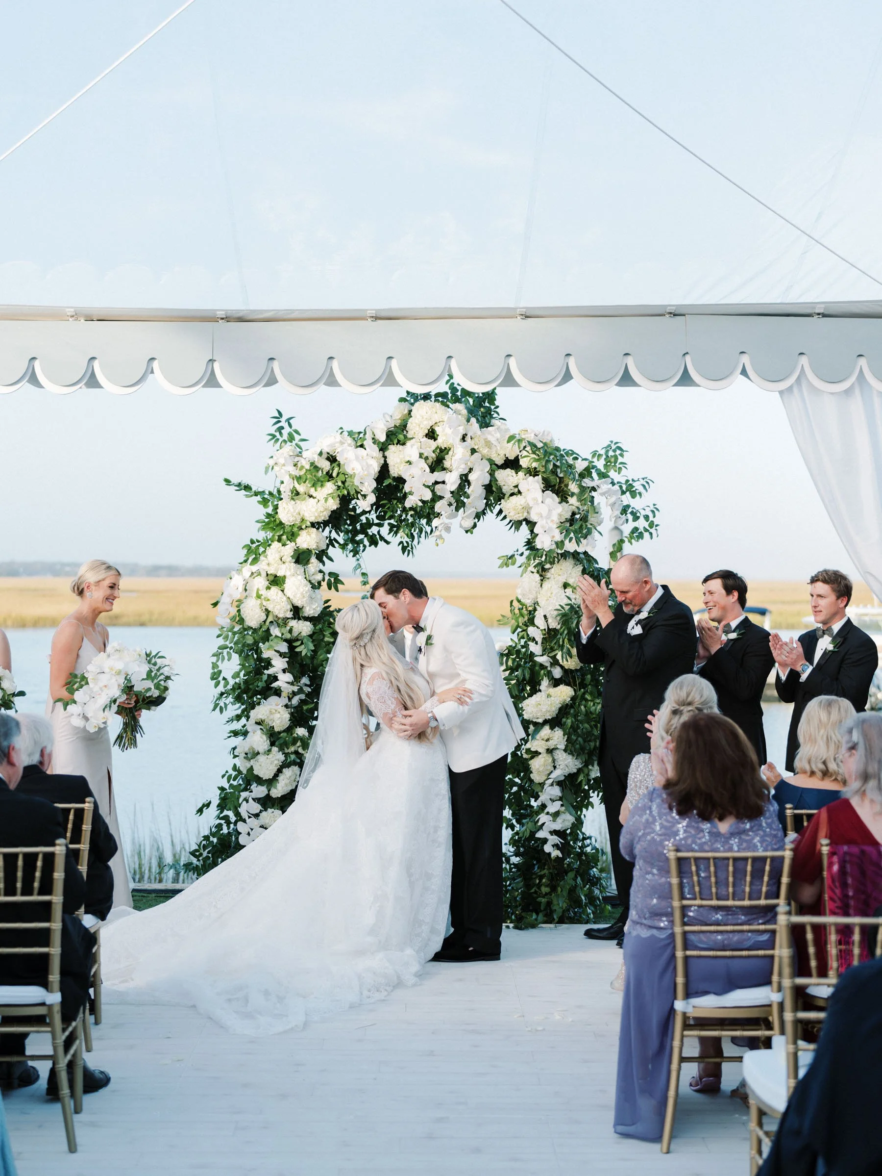 A wedding ceremony taking place outdoors under a white tent by a lake or river, with a bride and groom kissing under a floral arch made of white flowers and greenery, surrounded by bridesmaids and groomsmen, with guests seated watching.