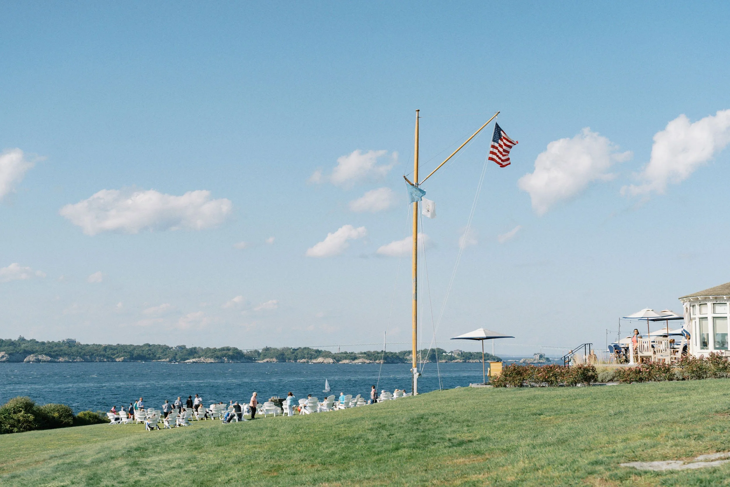 People sitting on white chairs on a grassy lawn by a body of water, with a flagpole flying an American flag and another flag, under a blue sky with scattered clouds, near a building with outdoor seating and umbrellas.