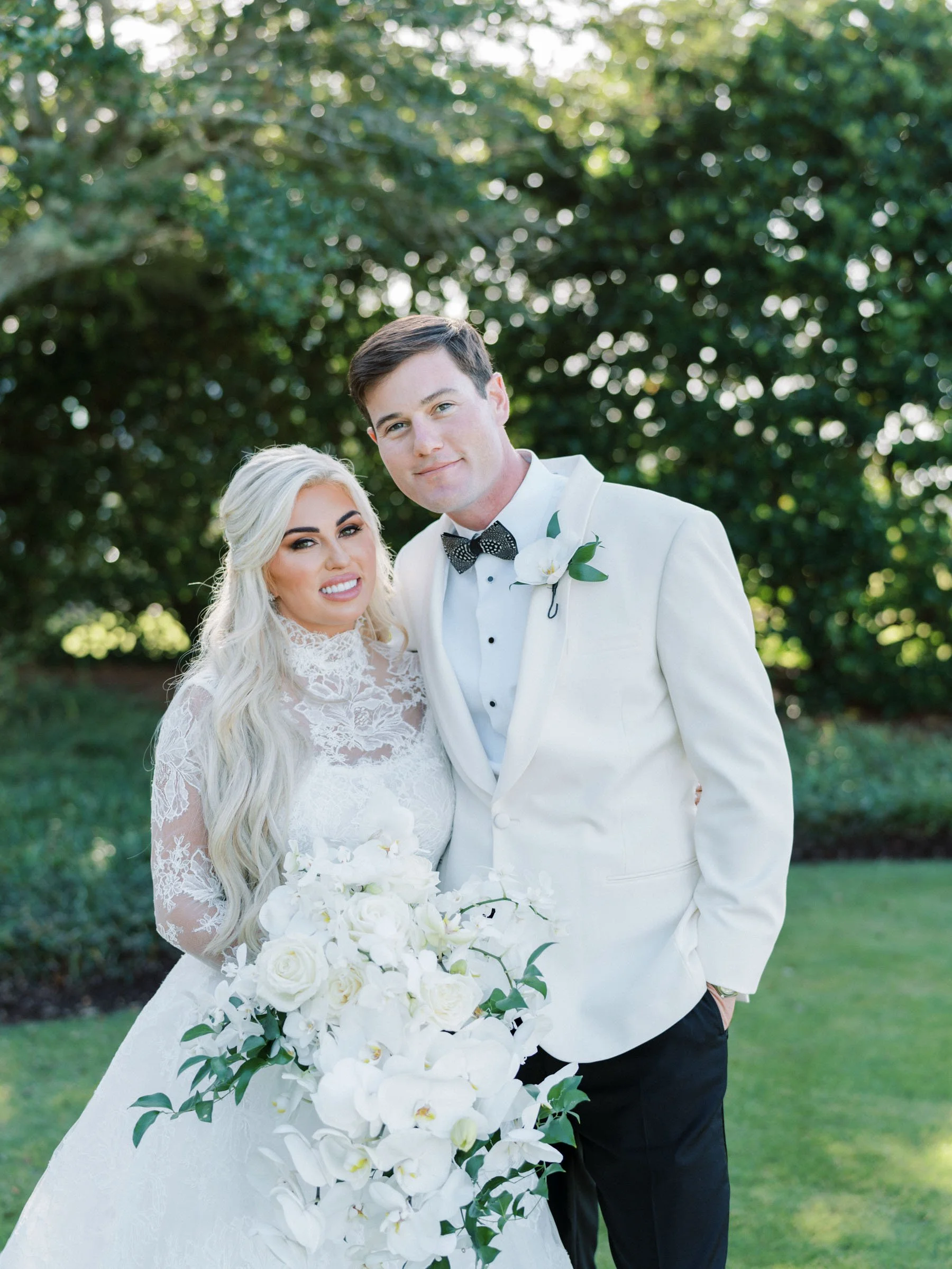 A bride with long, styled blonde hair in a white lace wedding dress holding a bouquet of white roses and orchids, standing next to a groom with dark hair in a white tuxedo jacket, black pants, and a black bow tie, outdoors with green trees in the bac