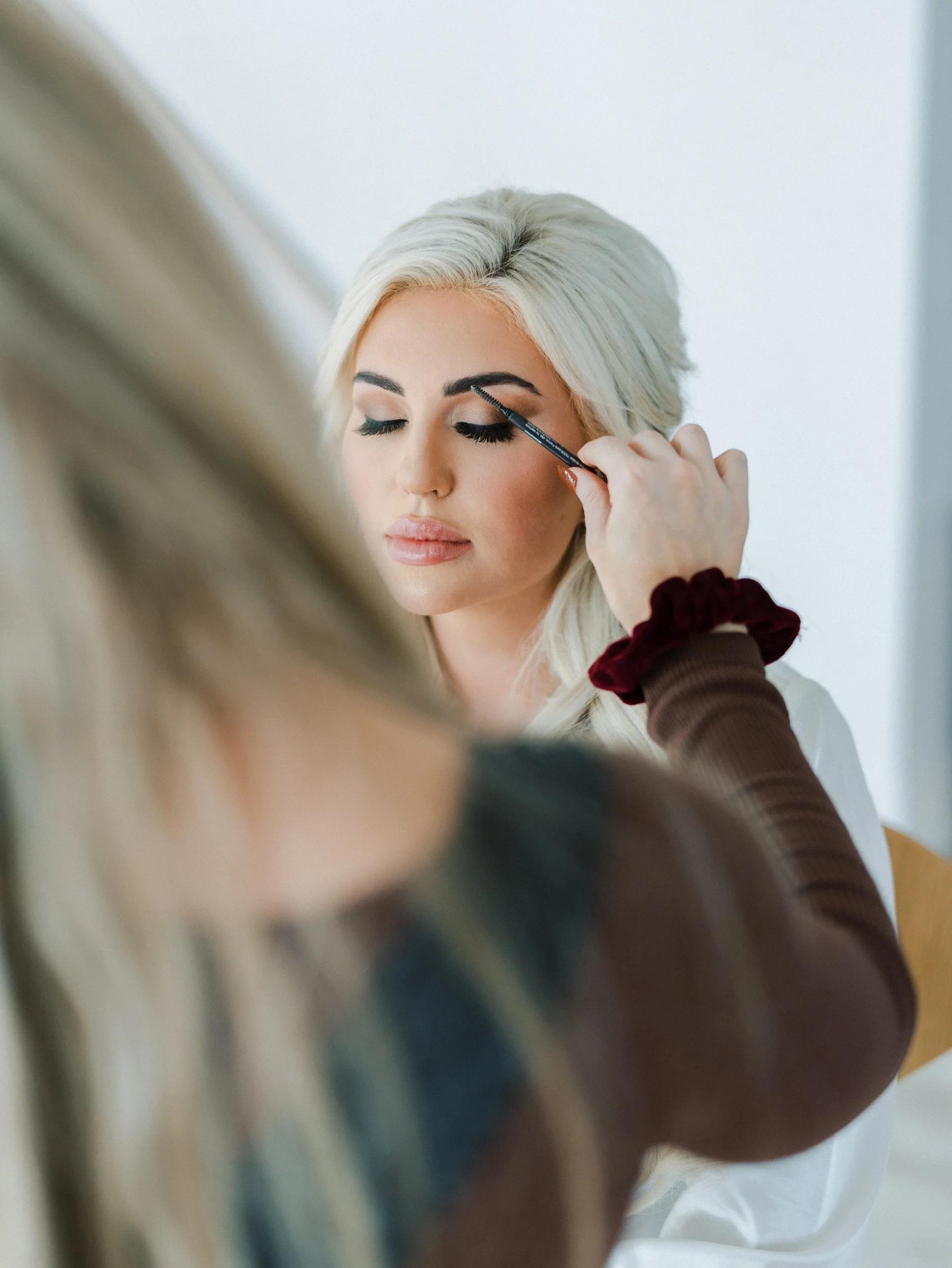 Woman with blonde hair getting makeup applied, eyes closed, by makeup artist.