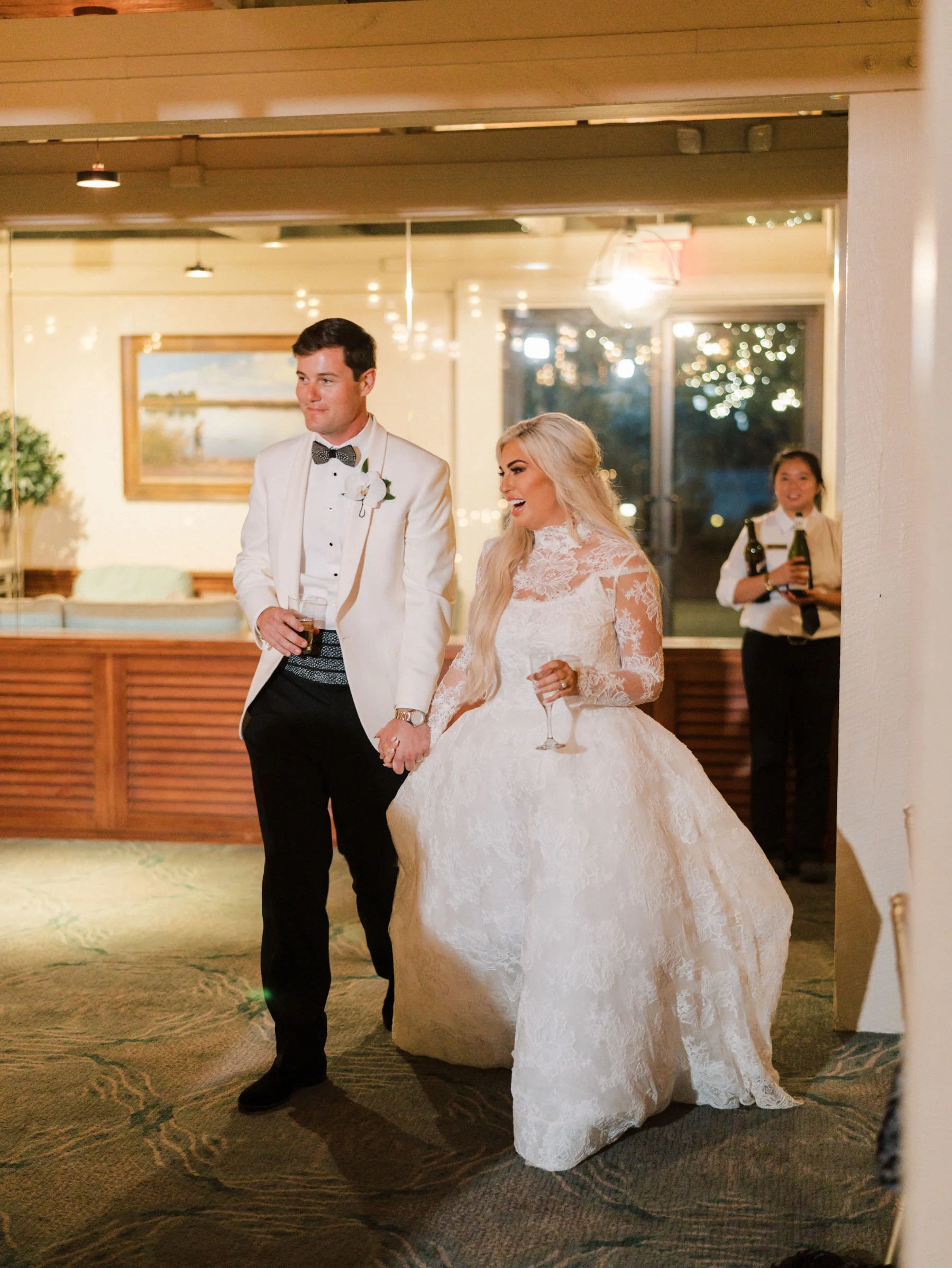 Bride and groom holding hands, walking at their wedding reception, smiling and dressed in formal wedding attire, with a server in the background holding drinks.