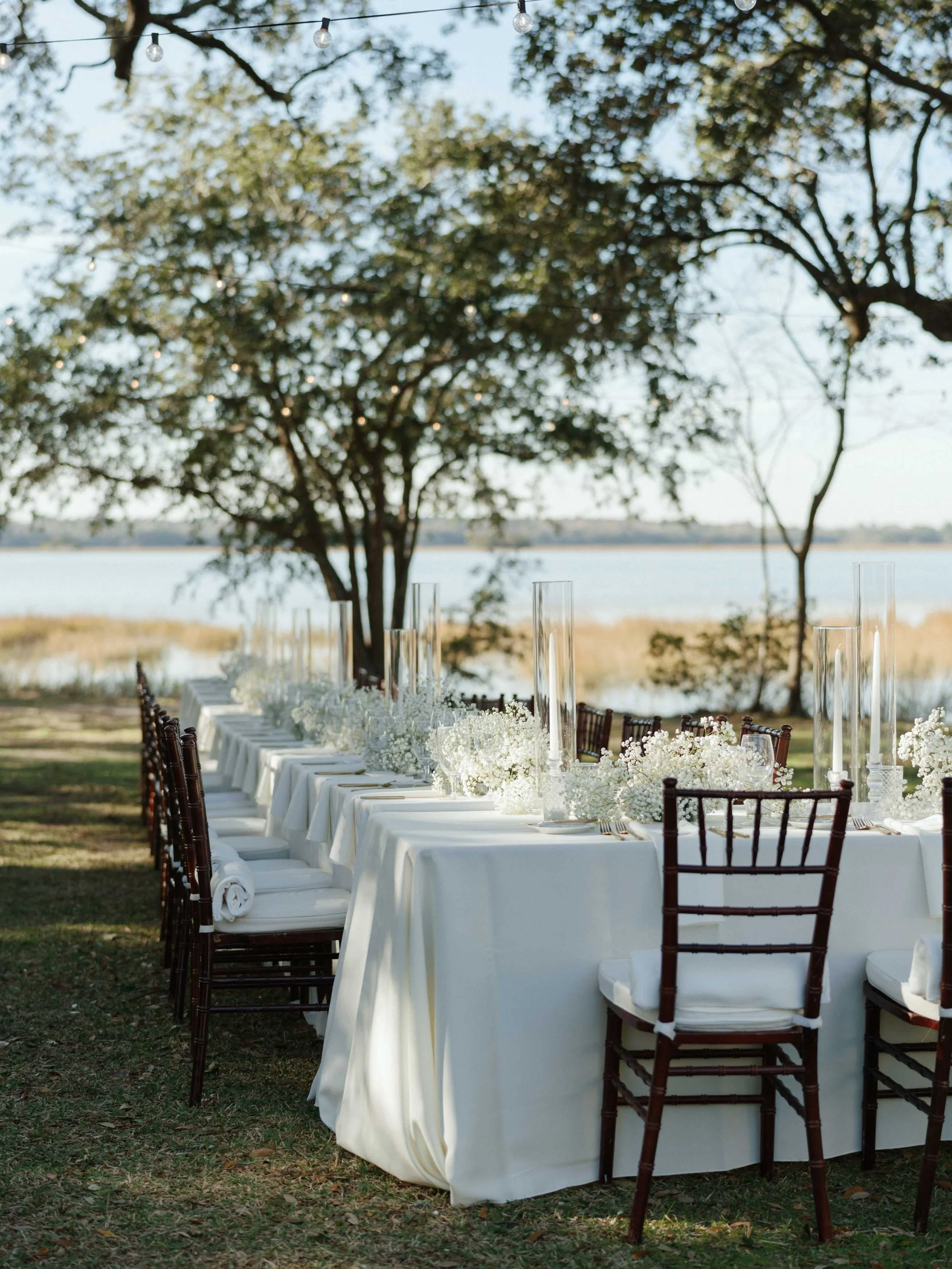 Outdoor wedding or event reception table set with white tablecloths, floral centerpieces, tall glass candle holders, and wooden chairs near a lake and trees.