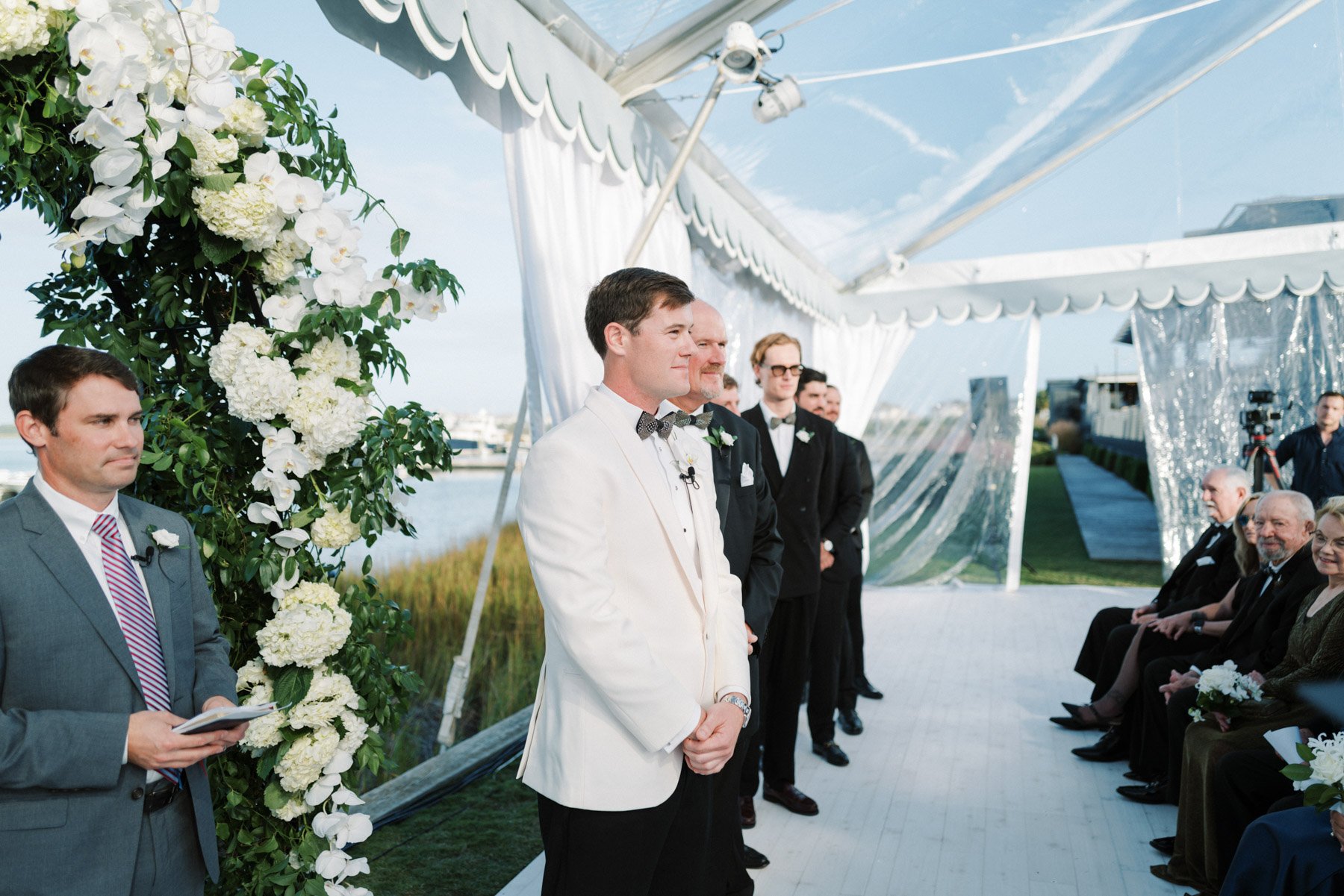 A wedding ceremony taking place outdoors under a white canopy decorated with white flowers, with groom and officiant at the front, and guests seated on the right side.