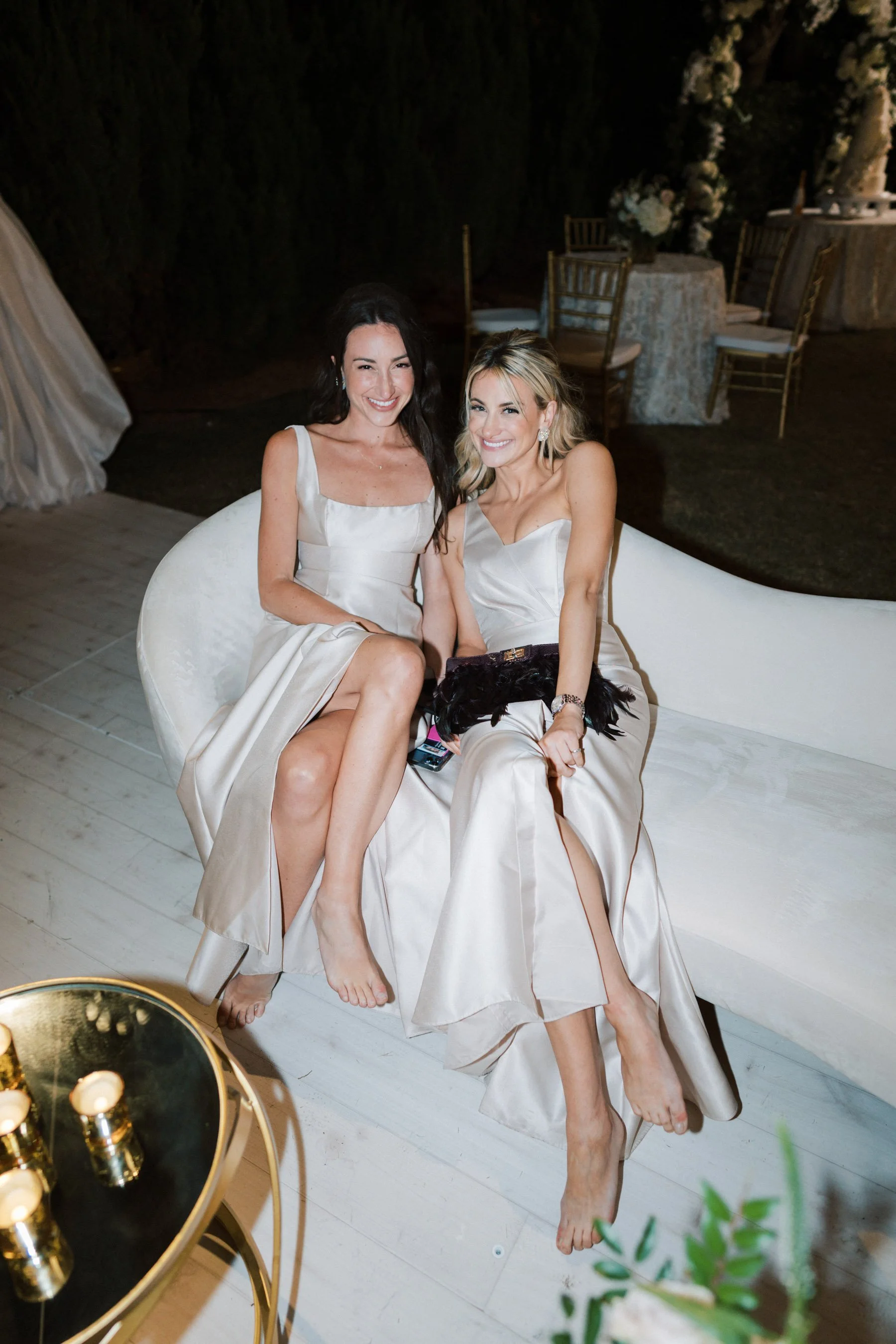Two women sitting on a cream-colored sofa at an outdoor event, smiling and barefoot, with tables and chairs in the background and a round table with candles in the foreground.