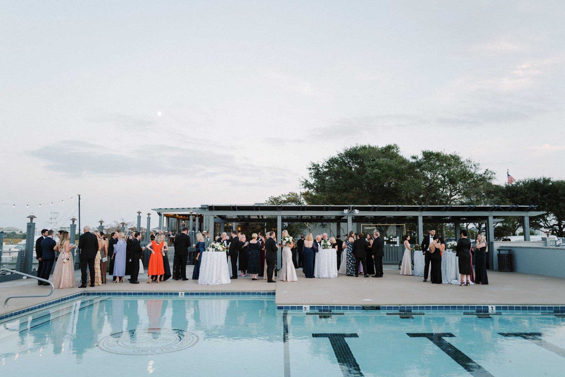 Guests gathered around standing tables with white tablecloths on a rooftop terrace during an outdoor event or wedding, with a pool in the foreground and trees and sky in the background.