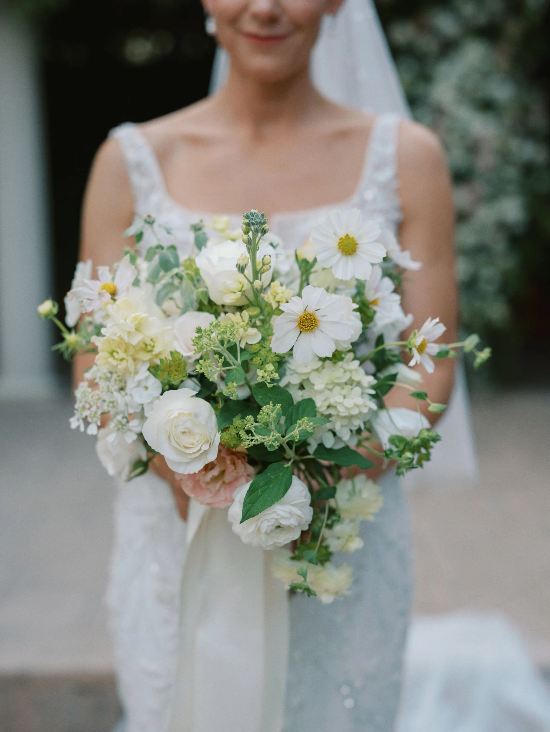 Bride holding a bouquet of white and pale pink flowers, with a blurred background.