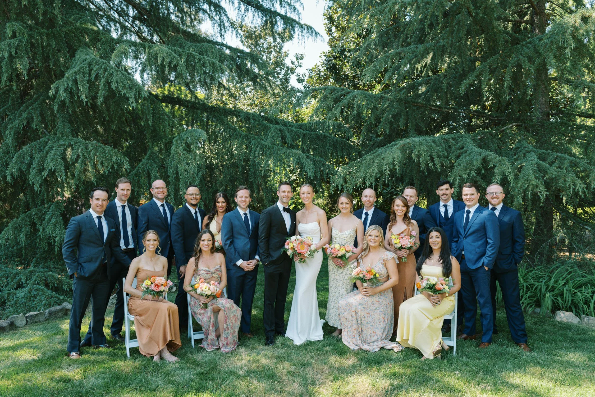 A group of people dressed in formal attire standing and sitting outdoors in front of green trees, at a wedding at Vanlandingham Estate