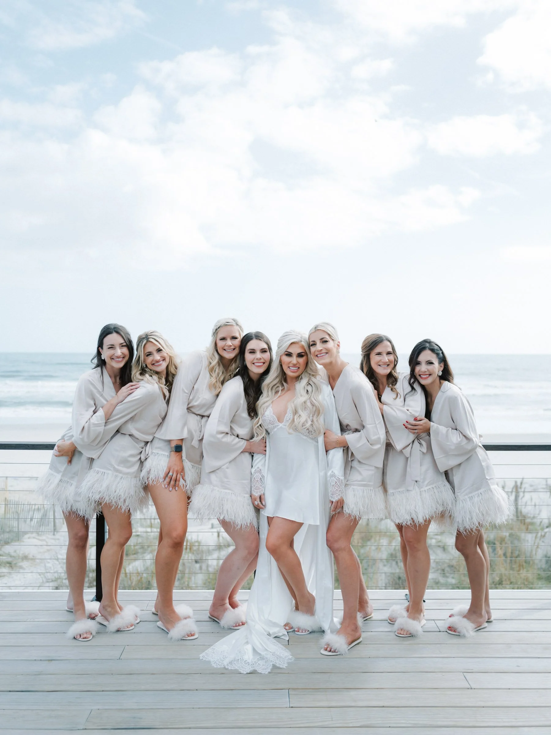 A group of women in white loungewear with feathered trim and fuzzy slippers, standing on a boardwalk by the beach, smiling for a photo.