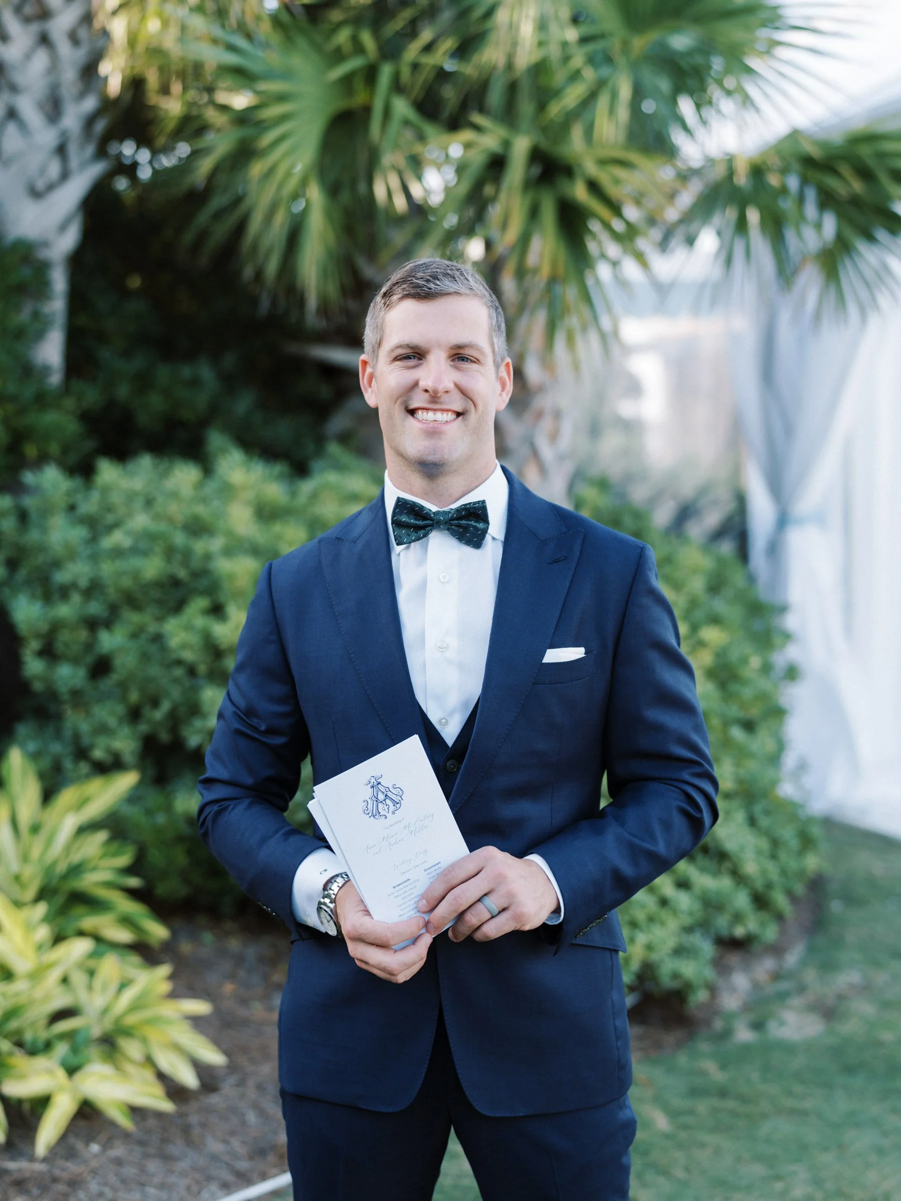 A man in a navy suit and bow tie holding a wedding invitation, smiling outdoors with green foliage and palm trees in the background.