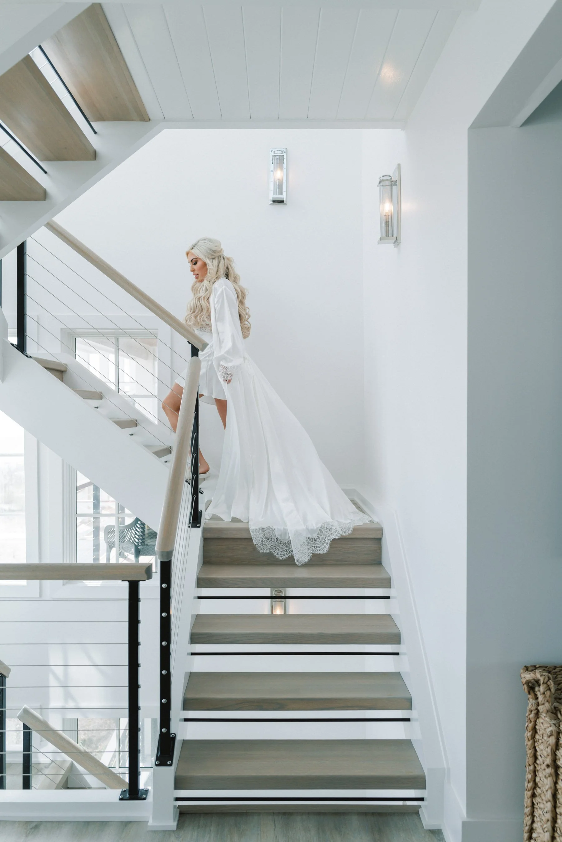 A woman with long, styled blonde hair in a white wedding gown ascending a staircase in a bright, modern interior.