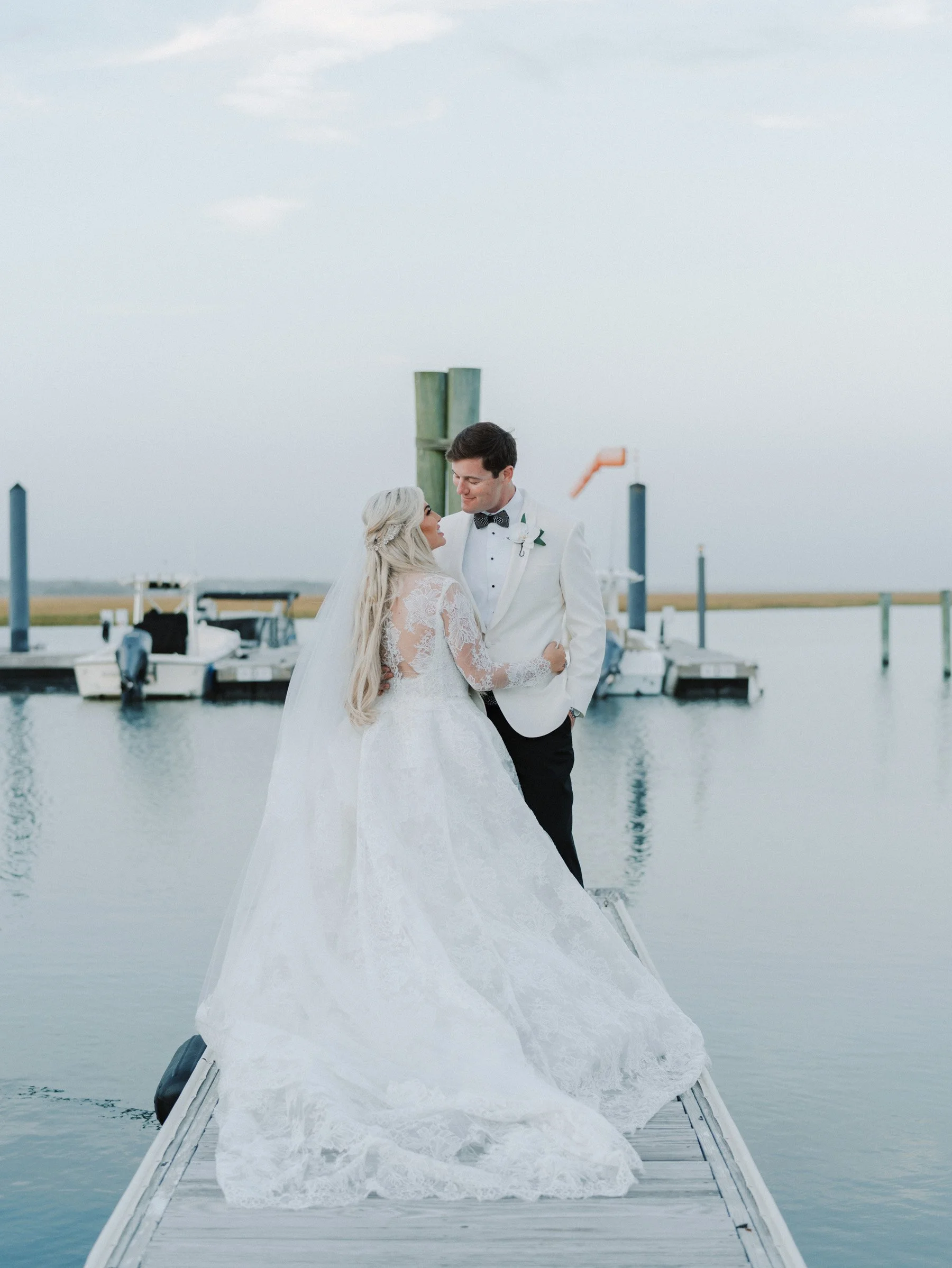 A bride and groom stand closely together on a dock by the water, gazing at each other, with a dock and boats in the background during a wedding photo shoot.