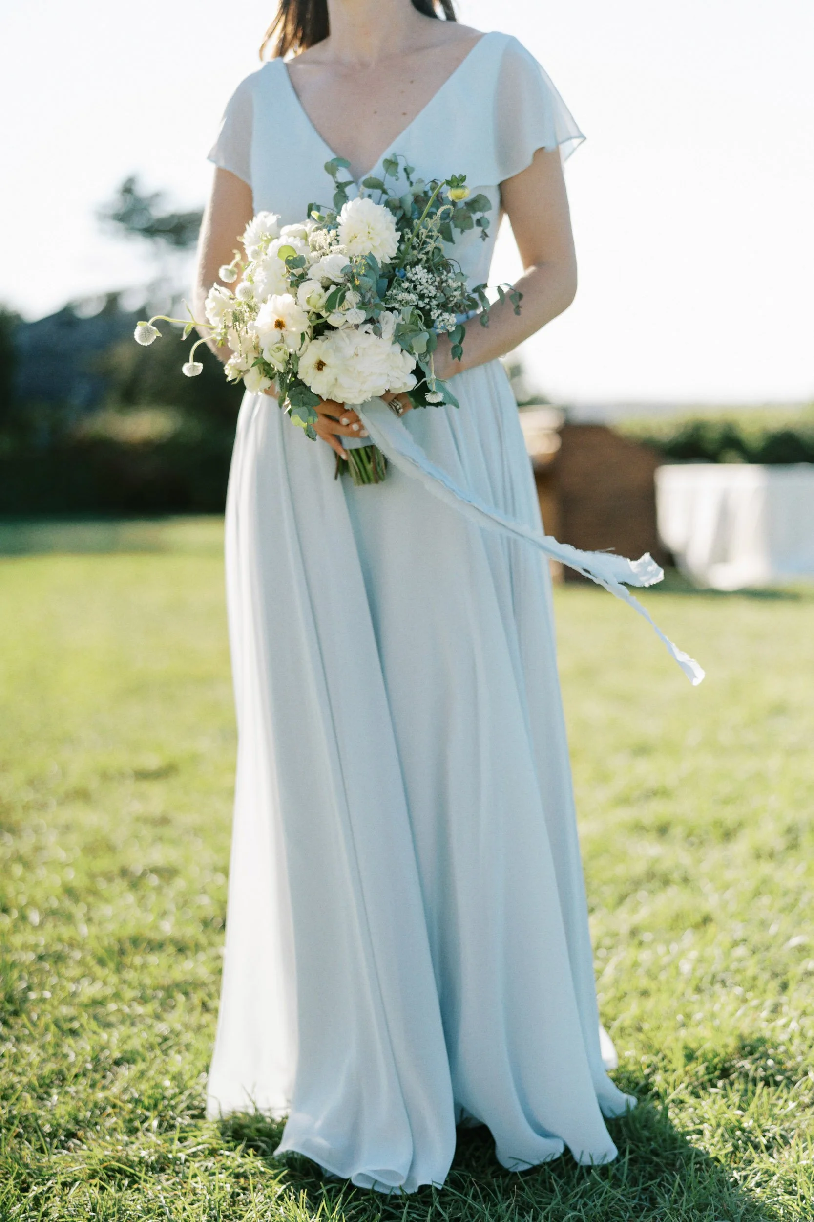 A woman dressed in a light blue gown holding a bouquet of white flowers outdoors on a sunny day.