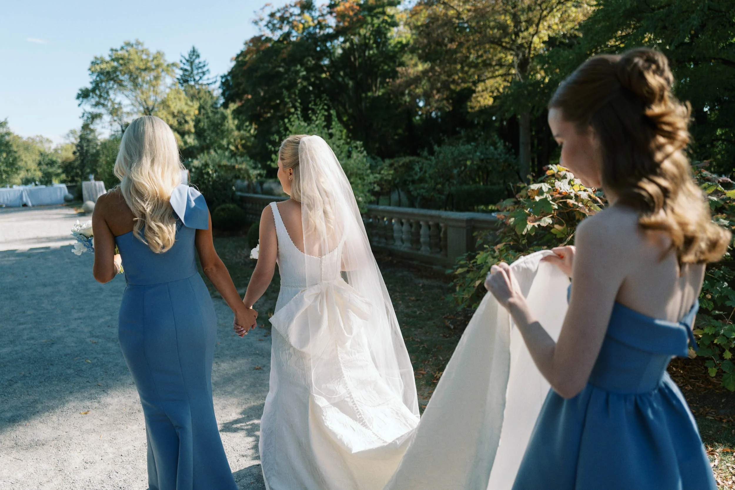 Three women, one in a wedding dress and two bridesmaids in blue dresses, walking outdoors on a sunny day holding hands, with trees and a decorative railing in the background.