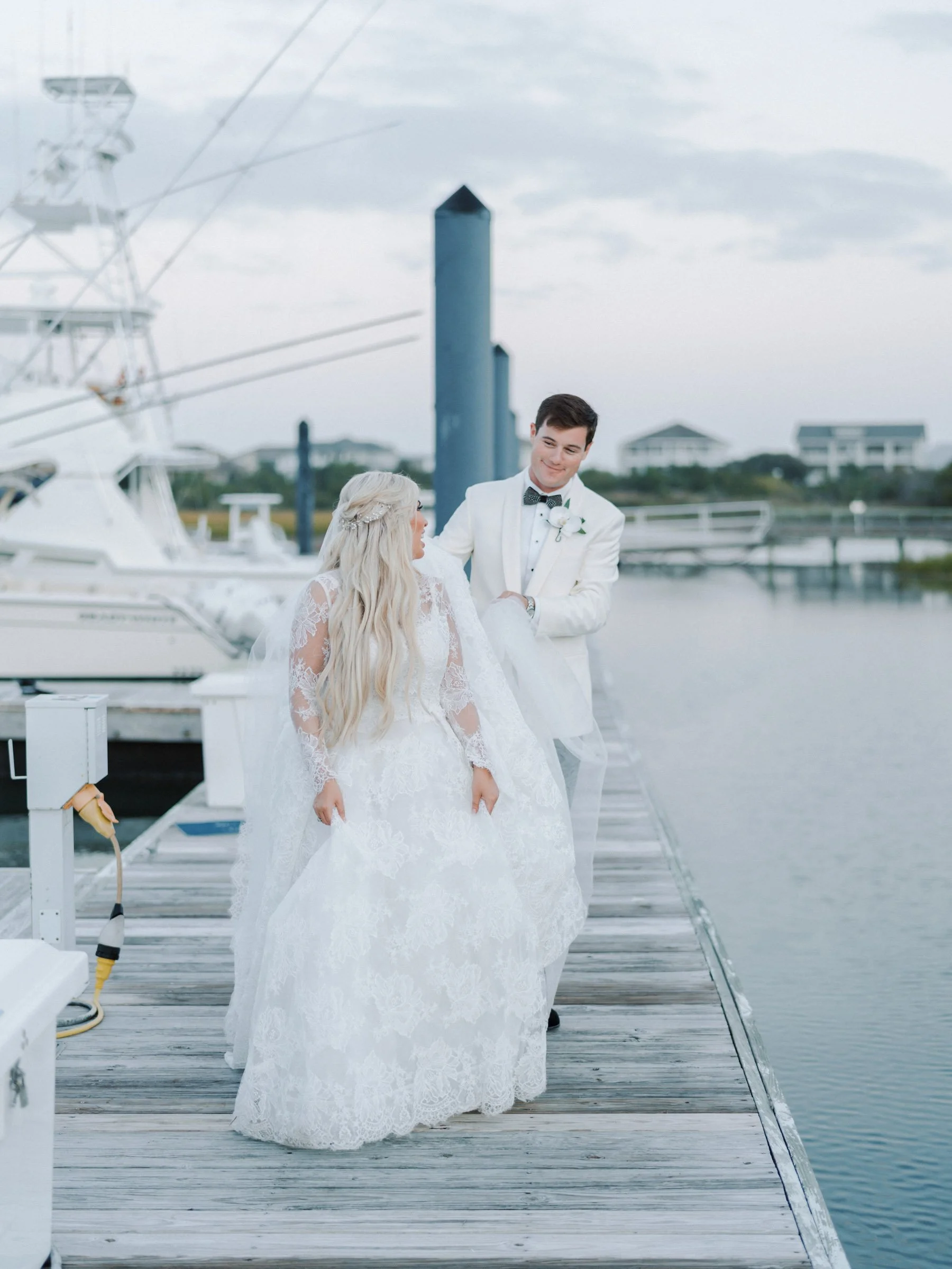 A bride and groom stand on a dock by the water, dressed in wedding attire, with boats and a marina in the background.