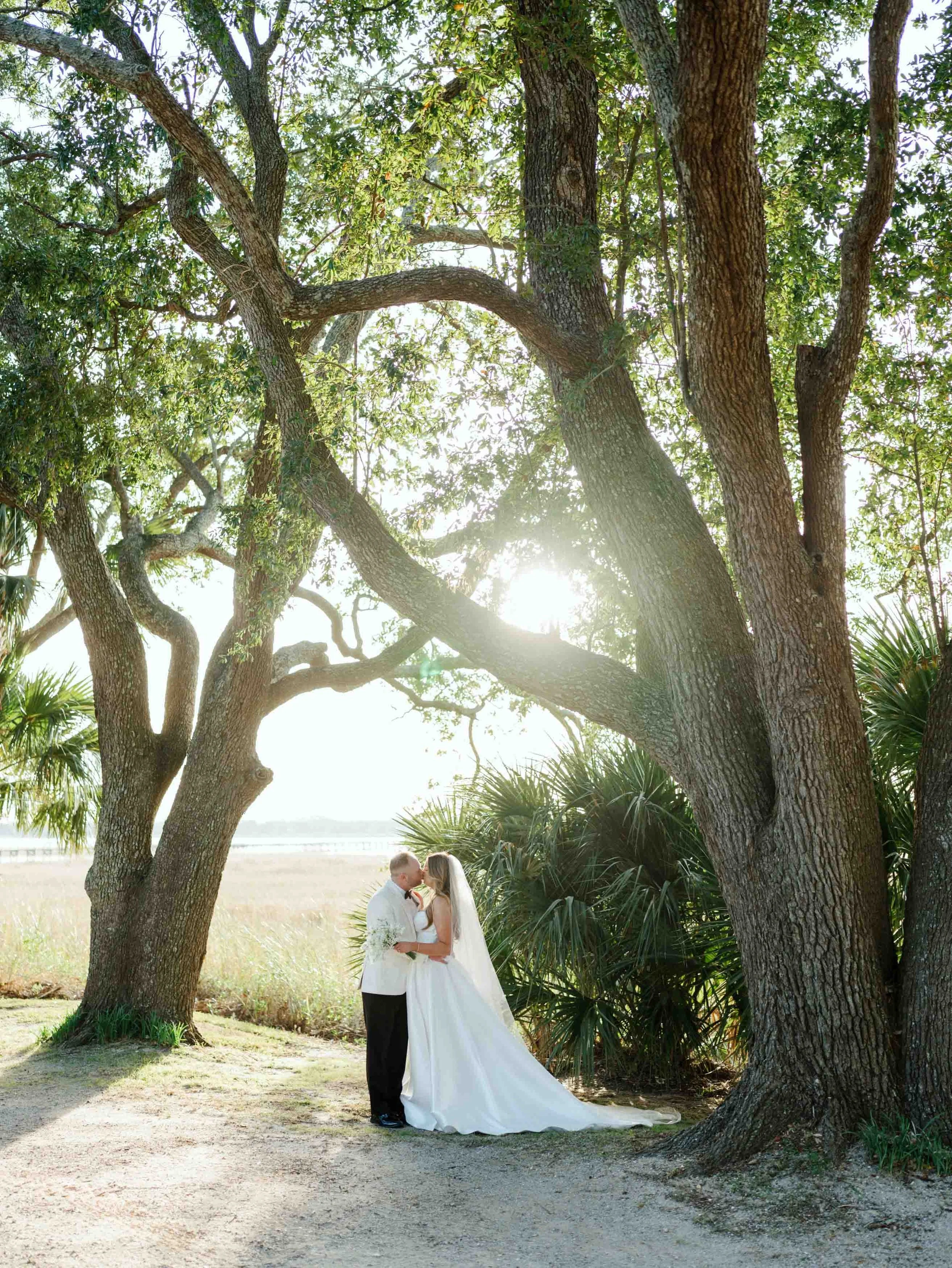 A bride and groom kissing beneath large trees with sunlight shining through the branches during their wedding.