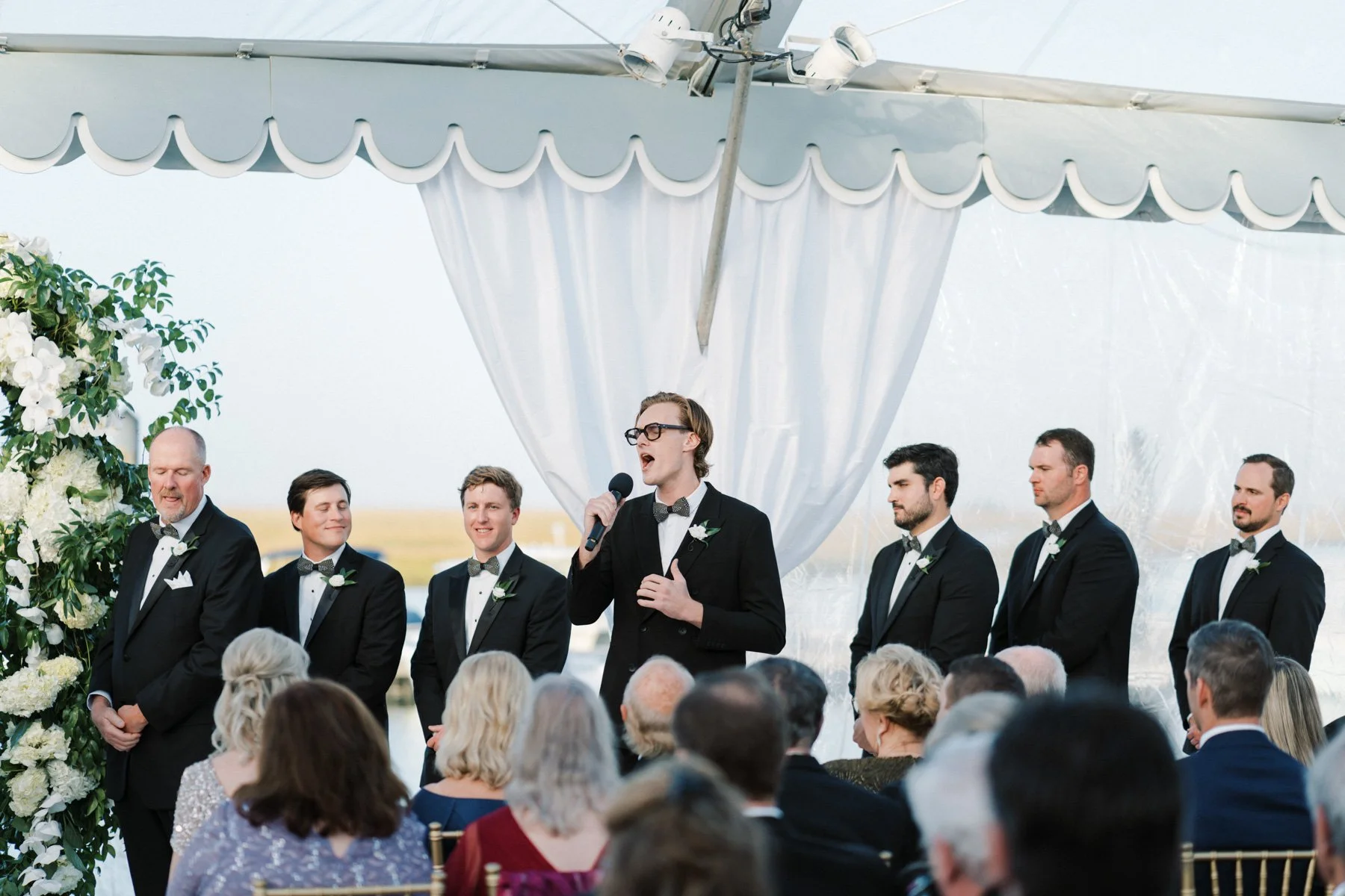 A groom and groomsmen standing during a wedding ceremony under a decorated white tent, with an audience seated in front.