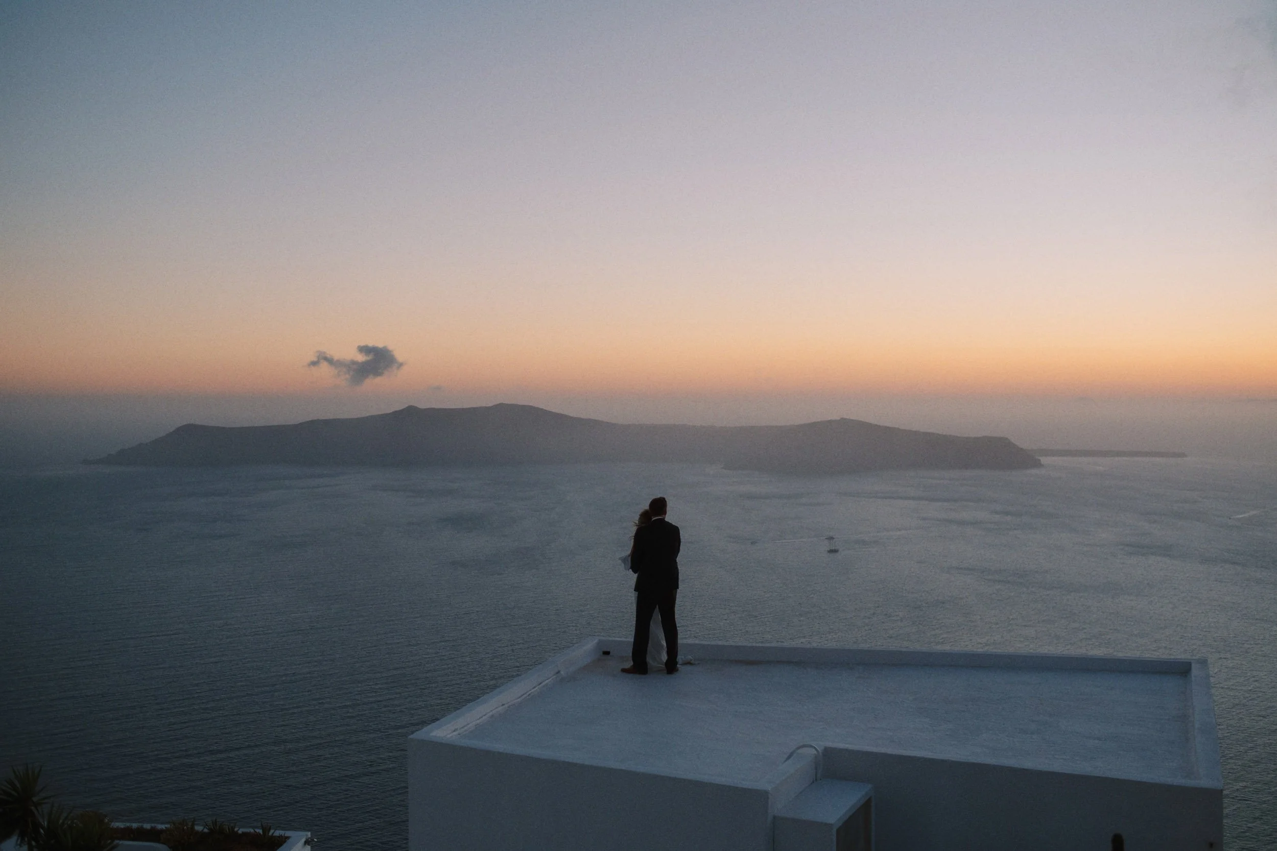 Person in formal attire standing on a white rooftop balcony, gazing at a sunset over the ocean with an island in the distance.