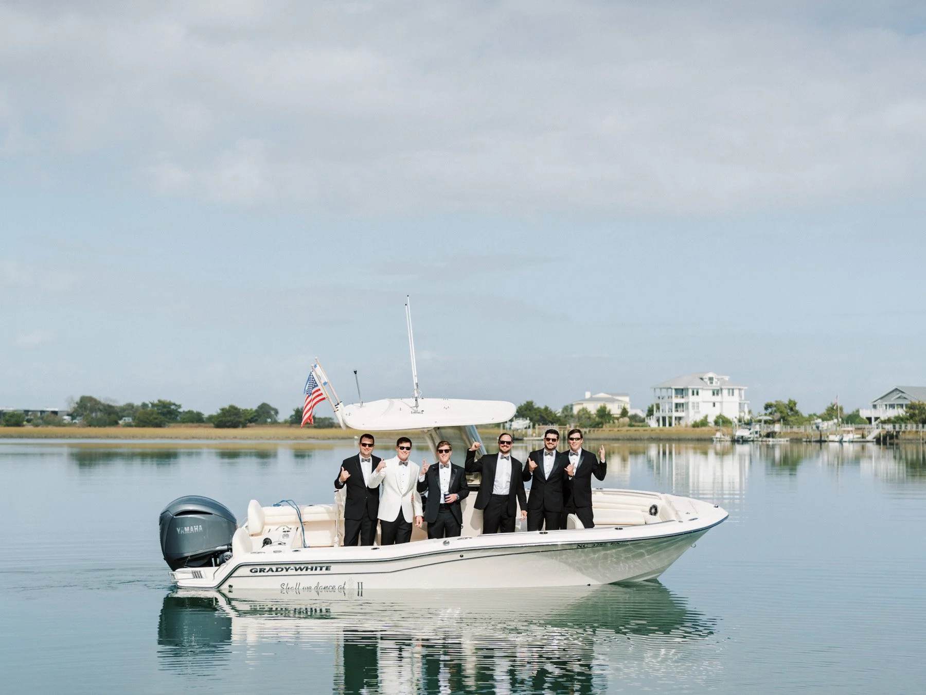 Group of six men in tuxedos on a boat in a calm waterway with houses and trees in the background.